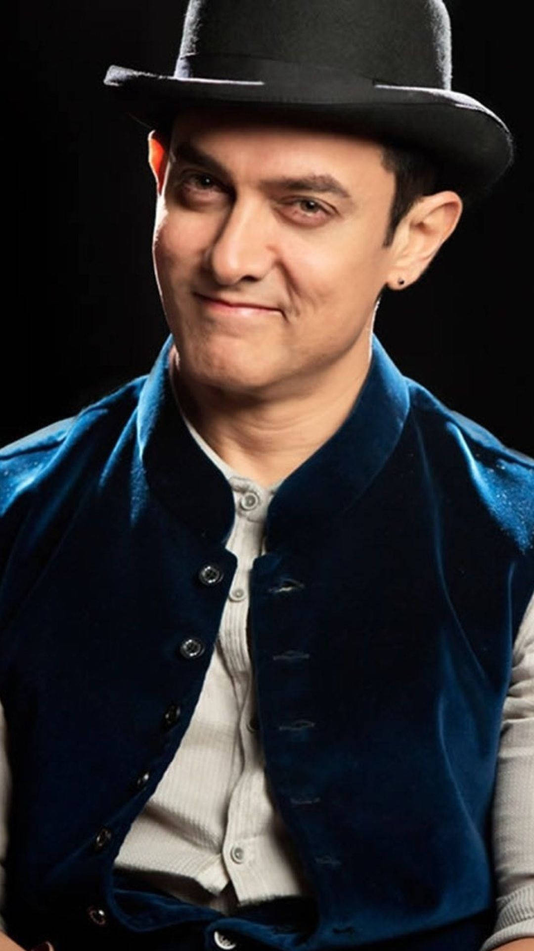 Aamir Khan 桌布 壁紙 — 一名戴黑色圓頂硬禮帽、穿藍色天鵝絨背心的男子在深色背景下微微微笑。