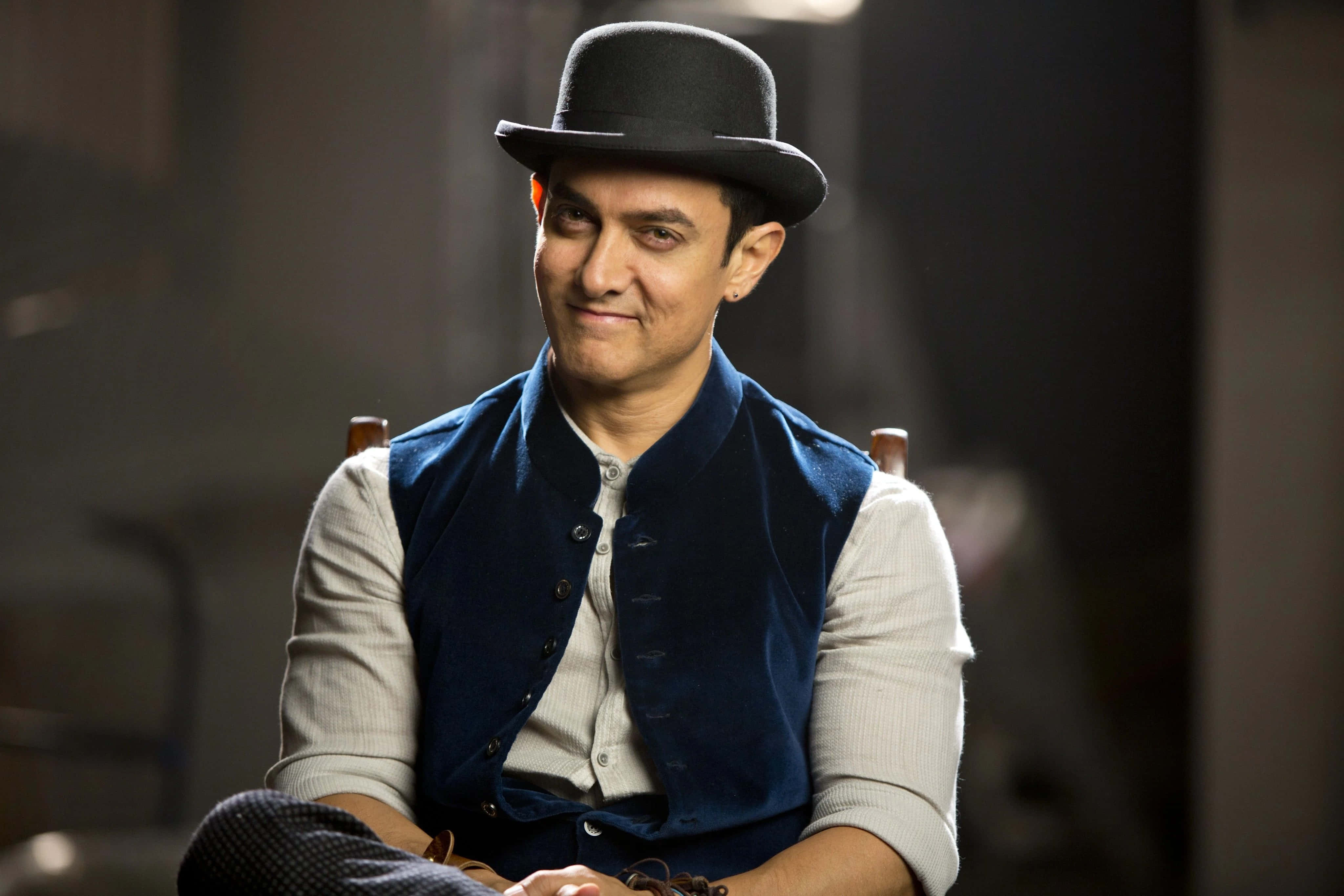 Aamir Khan 桌布 壁紙 — 一名戴黑色圓頂禮帽和藍色背心的男子坐在光線昏暗的房間裡，露出微妙的笑意。