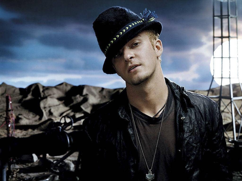 justin timberlake 桌布 壁紙 — 一名戴黑色帽子和皮夾克的男子站在戲劇性的多雲天空前，身後有一個大型燈光結構。