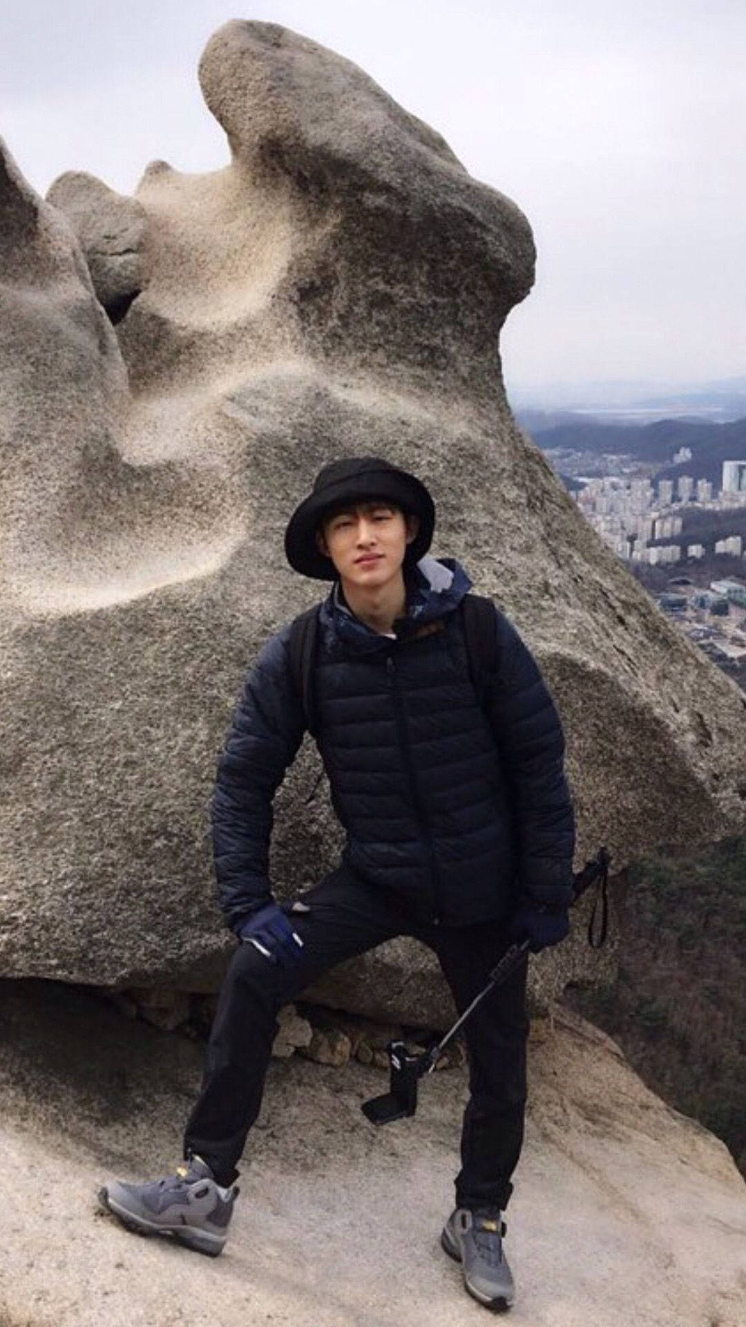 kim hanbin 桌布 壁紙 — 一名戴黑色帽子和穿羽絨外套的年輕男子自信地站在一個巨大的岩石構造旁，俯瞰著陰天下一片廣闊的城市景觀。