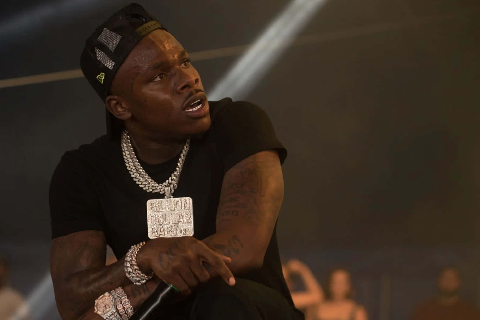 Dababy 桌布 壁紙 — 一名戴黑色帽子和穿黑色襯衫的男子，坐在舞台燈光下，戴著一條帶有「BILLION DOLLAR」吊墜的大銀鏈。