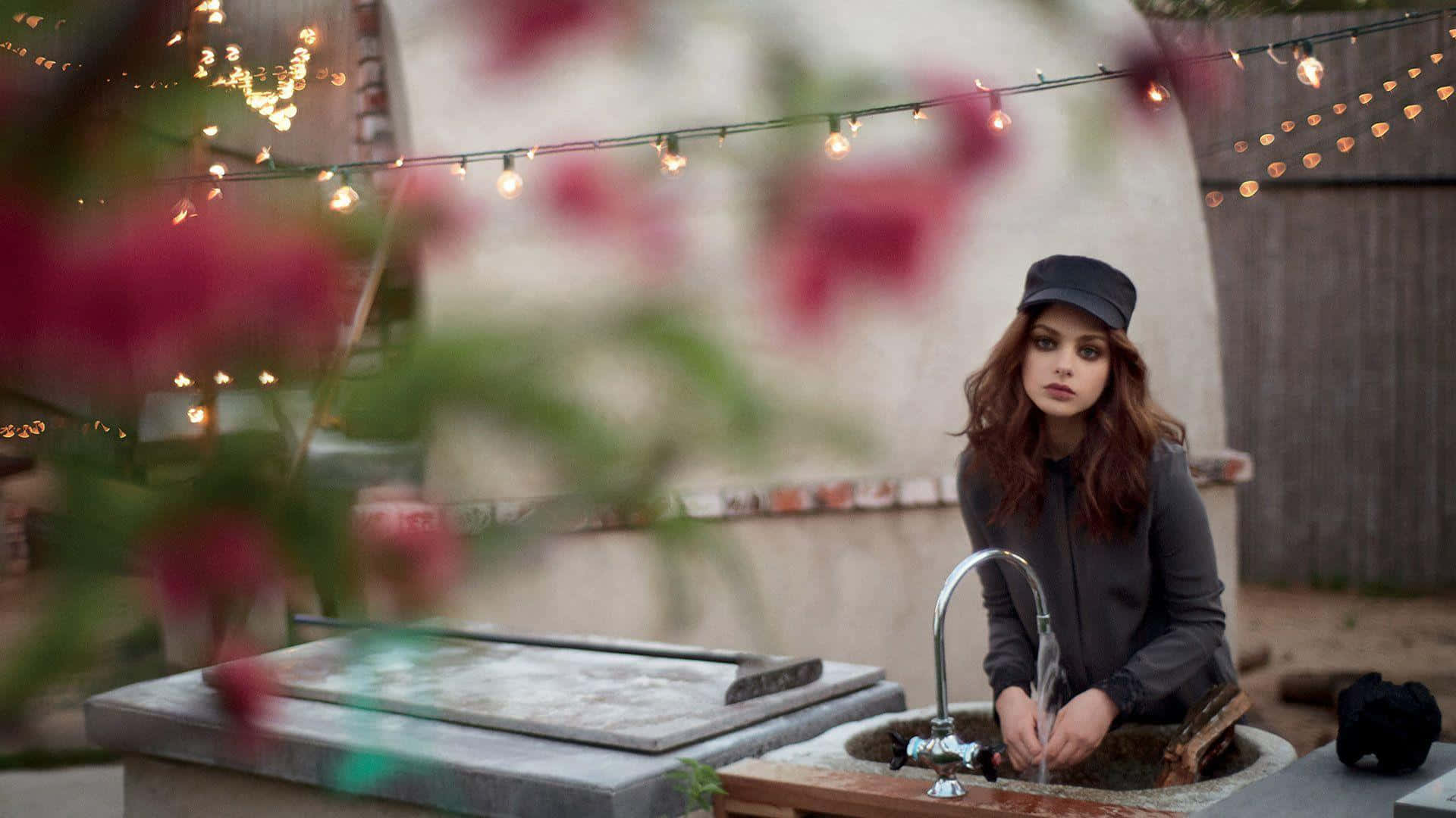 Odeya Rush 桌布 壁紙 — 一名戴黑色帽子的女性在戶外洗手台洗手，上方有燈串，前景有模糊的粉紅色花朵。
