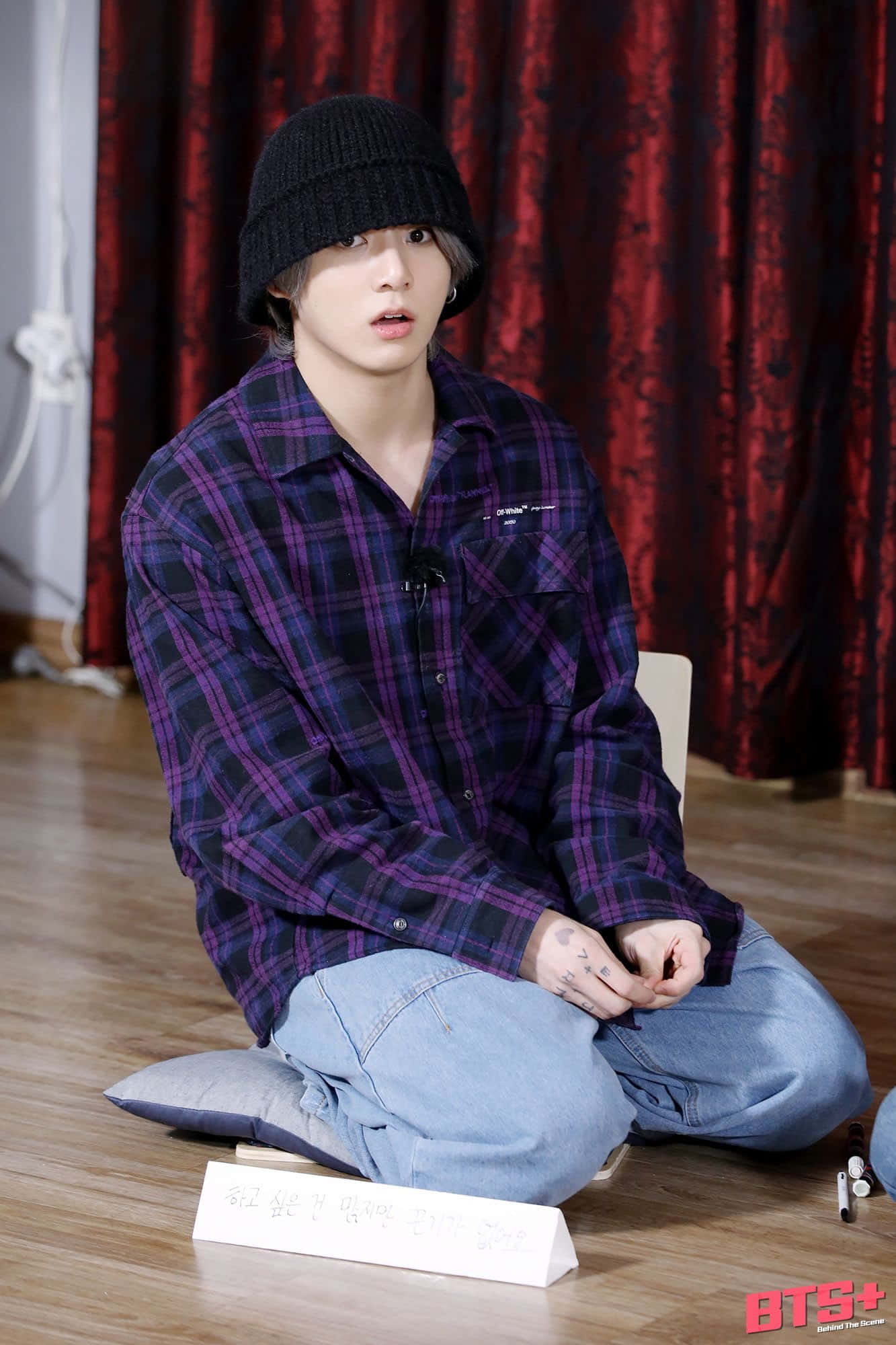 Jungkook 桌布 壁紙 — 一名戴黑色毛帽、身穿紫色格紋襯衫的年輕男子，盤腿坐在紅色天鵝絨窗簾前的木地板上。