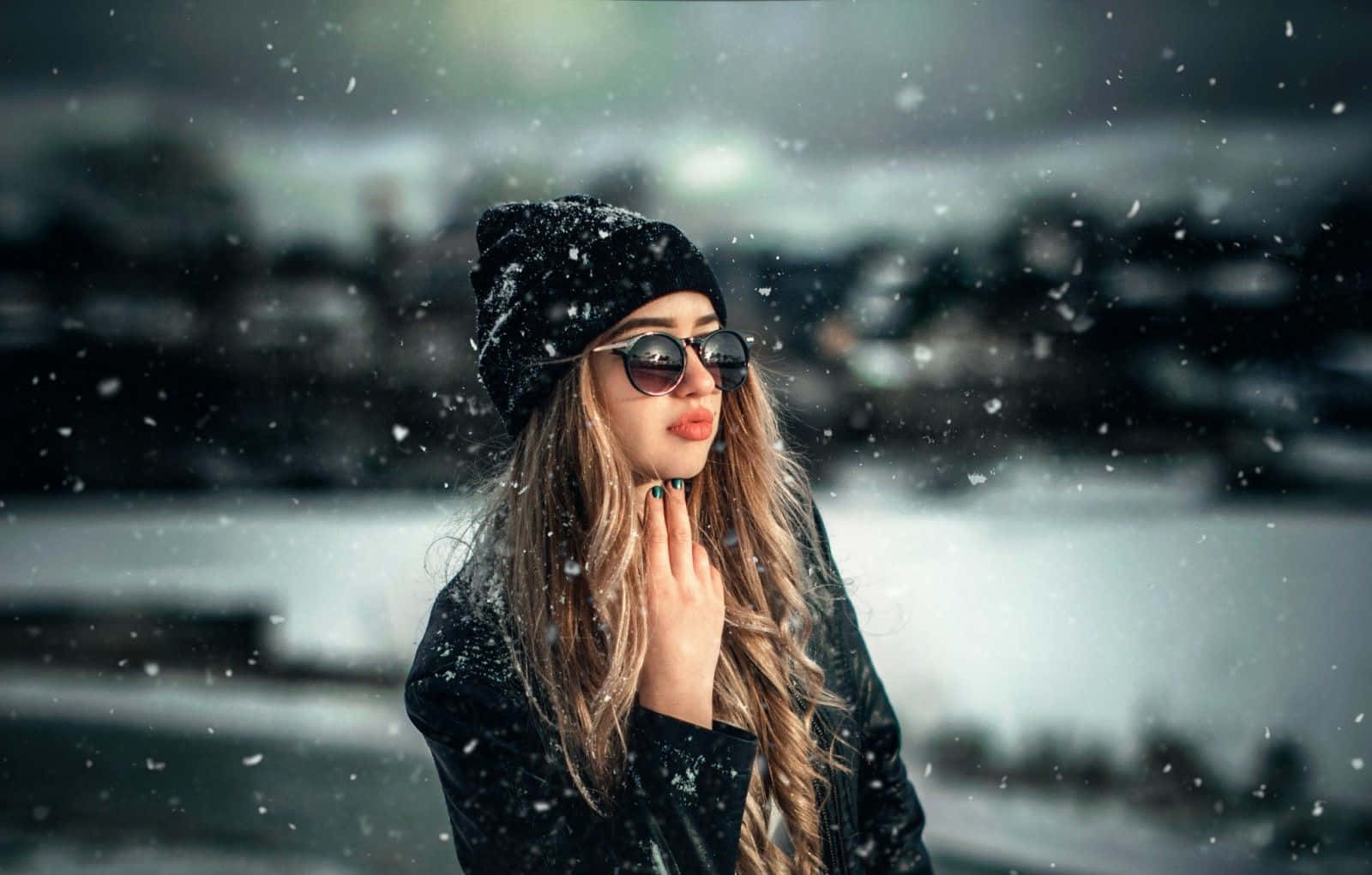Winter Fashion 桌布 壁紙 — 一名戴黑色毛帽和太陽眼鏡的女子站在飄雪中，背後是模糊的海濱城市景觀。