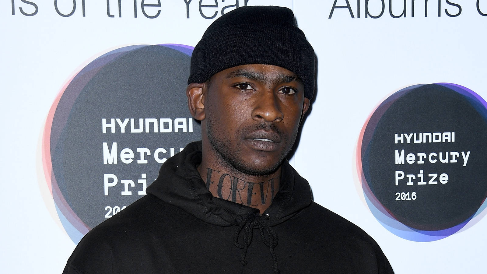 Skepta 桌布 壁紙 — 一名戴黑色毛帽和連帽上衣的男子站在Hyundai Mercury Prize活動現場，身後有標誌。