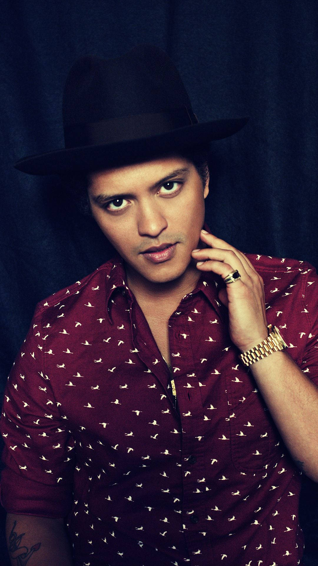 Bruno Mars 桌布 壁紙 — 一名戴黑色軟呢帽、穿紅色圖案襯衫的男子，在黑暗背景下將手靠近臉部擺姿勢。