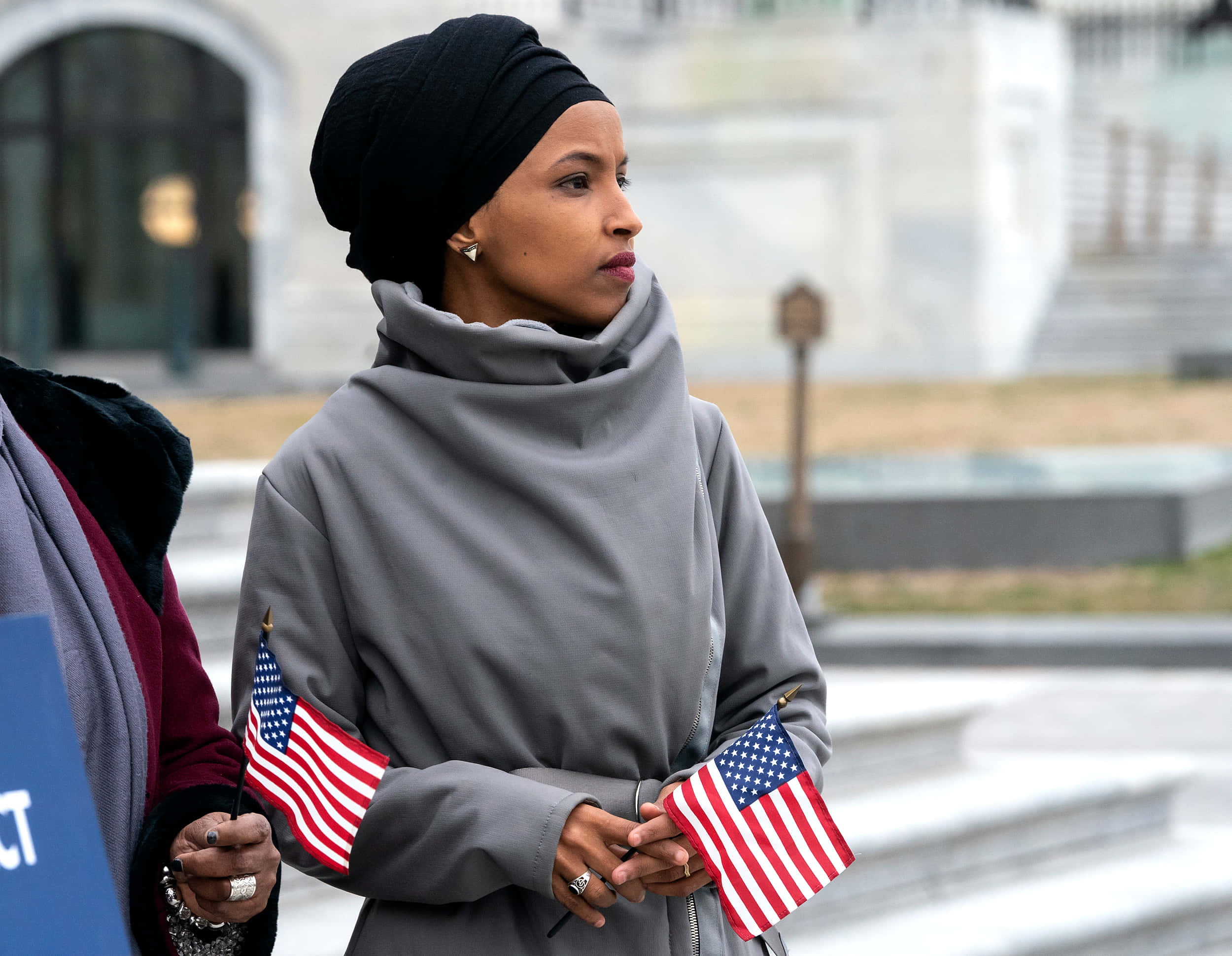 USA Flag 桌布 壁紙 — 一名戴黑色頭巾、穿灰色外套的女性，在政府大樓附近的戶外手持兩面美國國旗。