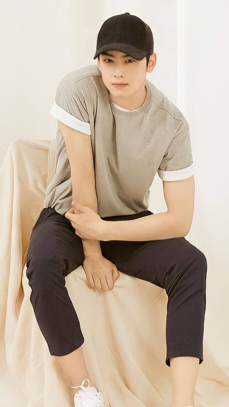 Eunwoo 桌布 壁紙 — 一名戴黑色鴨舌帽、穿灰色T恤的年輕男子，隨意地坐在一張米色布藝椅上，背景是柔和的白色。