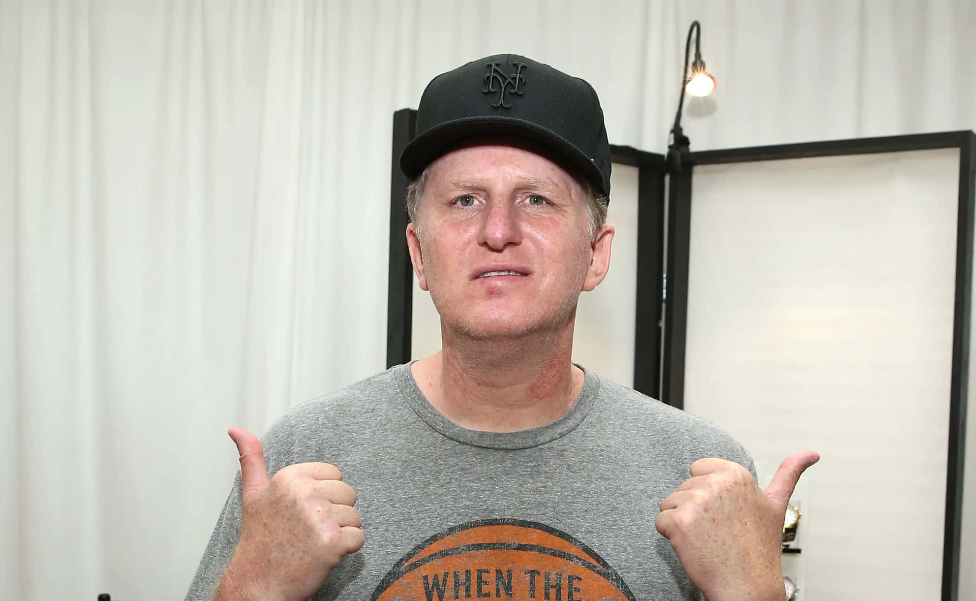 Michael Rapaport 桌布 壁紙 — 一名戴黑色鴨舌帽、穿灰色T恤的男子，在白色窗簾背景和一個懸掛燈泡前豎起兩個大拇指。