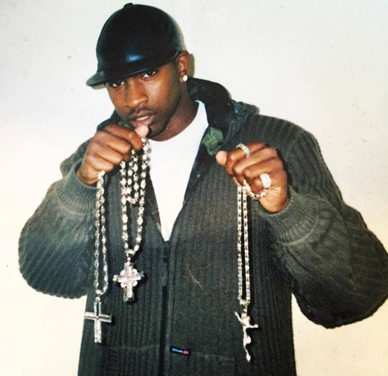 Skepta 桌布 壁紙 — 一名戴黑色鴨舌帽、穿綠色毛衣的男子，在素色背景前手持兩條帶十字架吊墜的華麗銀色玫瑰經念珠。