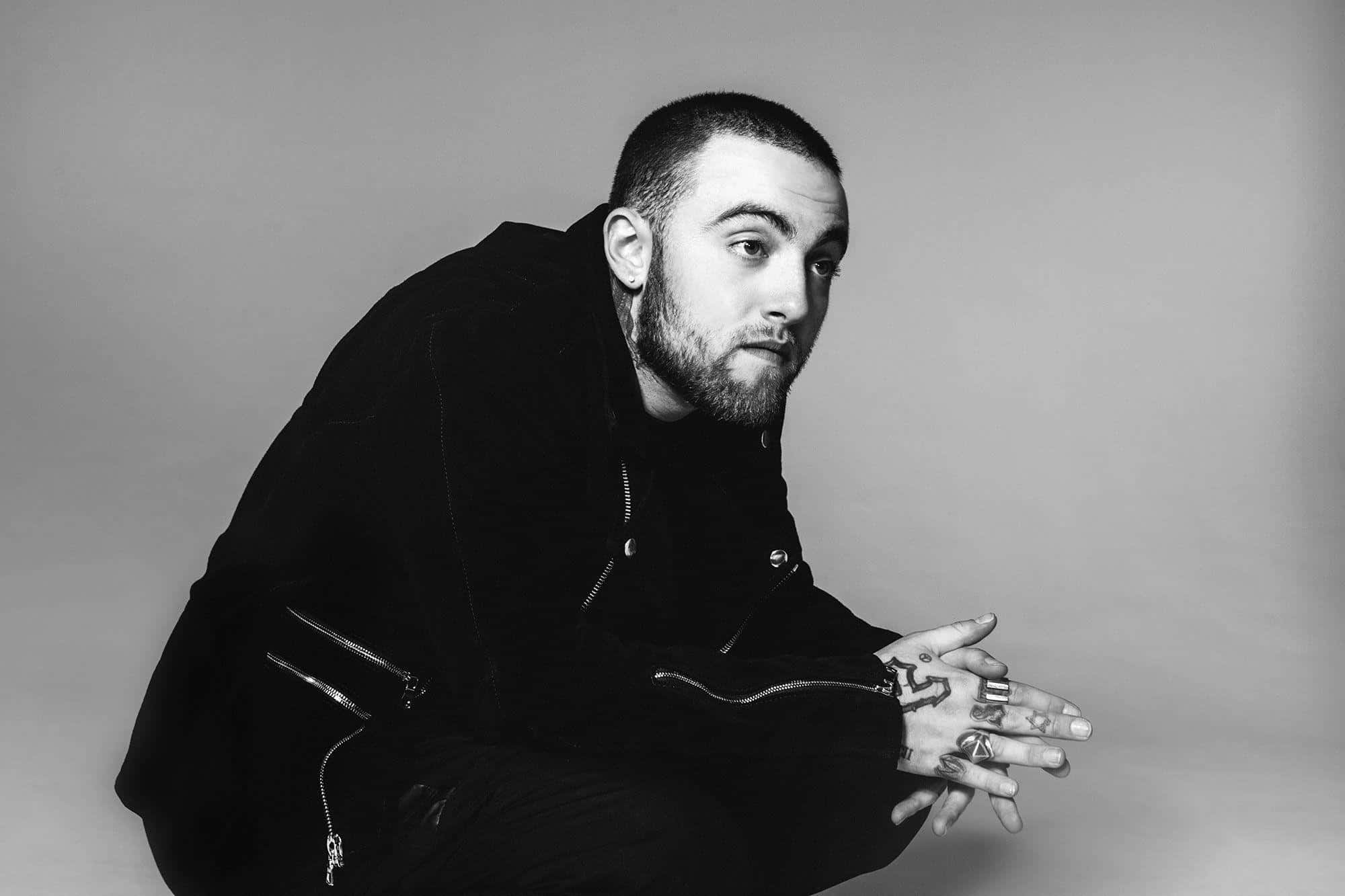 Mac Miller 桌布 壁紙 — 一名手上有刺青的男子穿著黑色衣服，在單色調的純灰色背景前蹲下。
