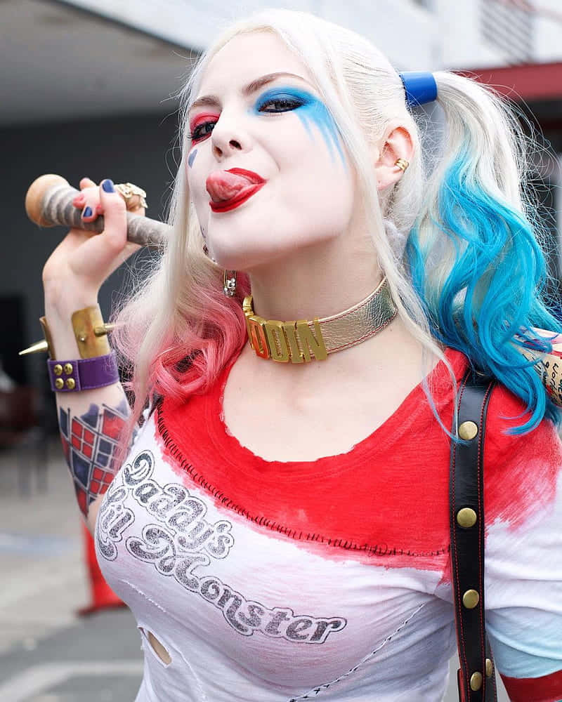 Harley Quinn Cosplay 桌布 壁紙 — 一名打扮成哈莉·奎茵的女性在都市環境中伸出舌頭，同時手持棒球棍。