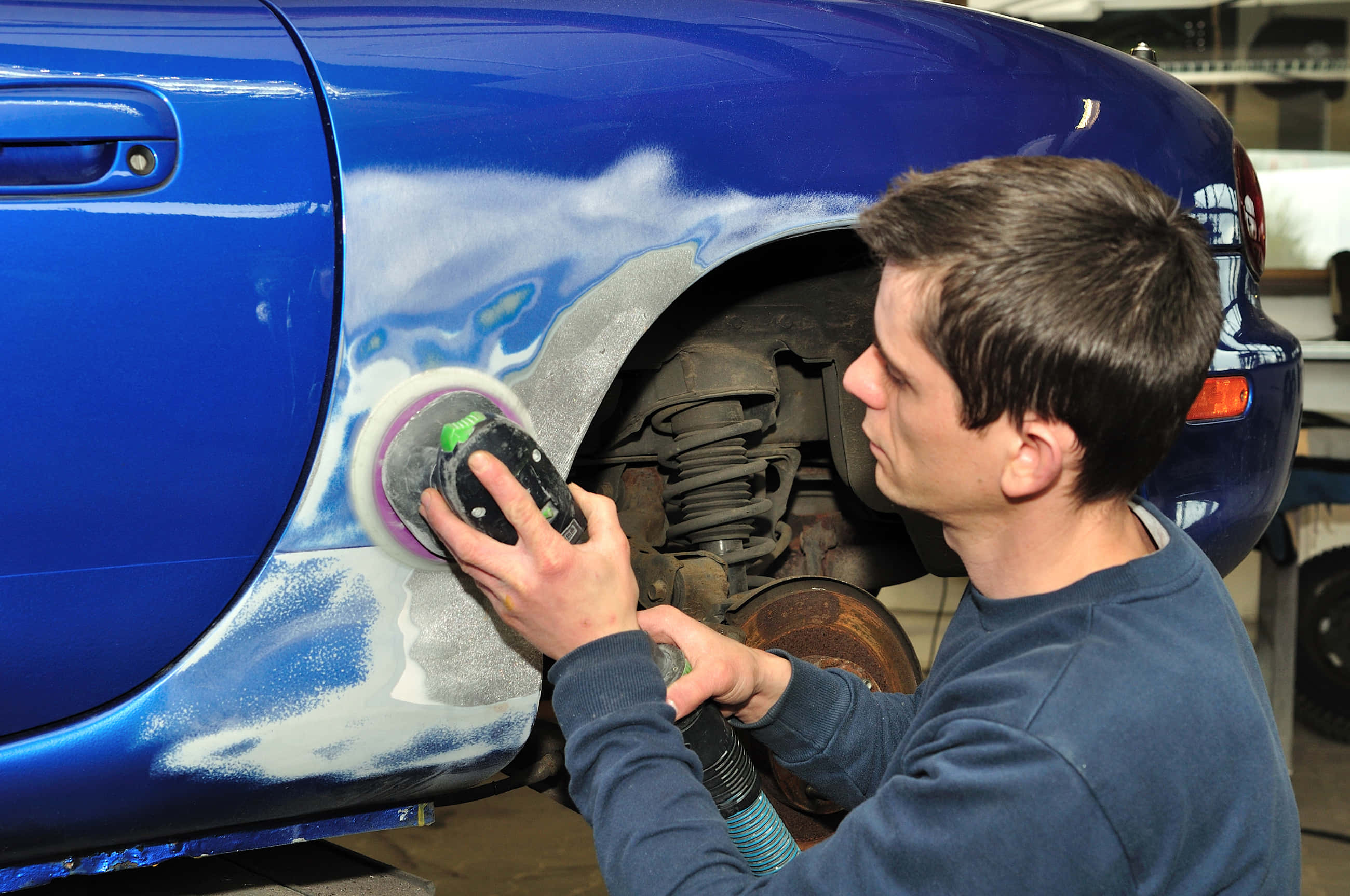 Car Repair 桌布 壁紙 — 一名技師在光線充足的車庫中，拋光一輛鮮藍色汽車的側面，泡沫和灰塵飛揚。