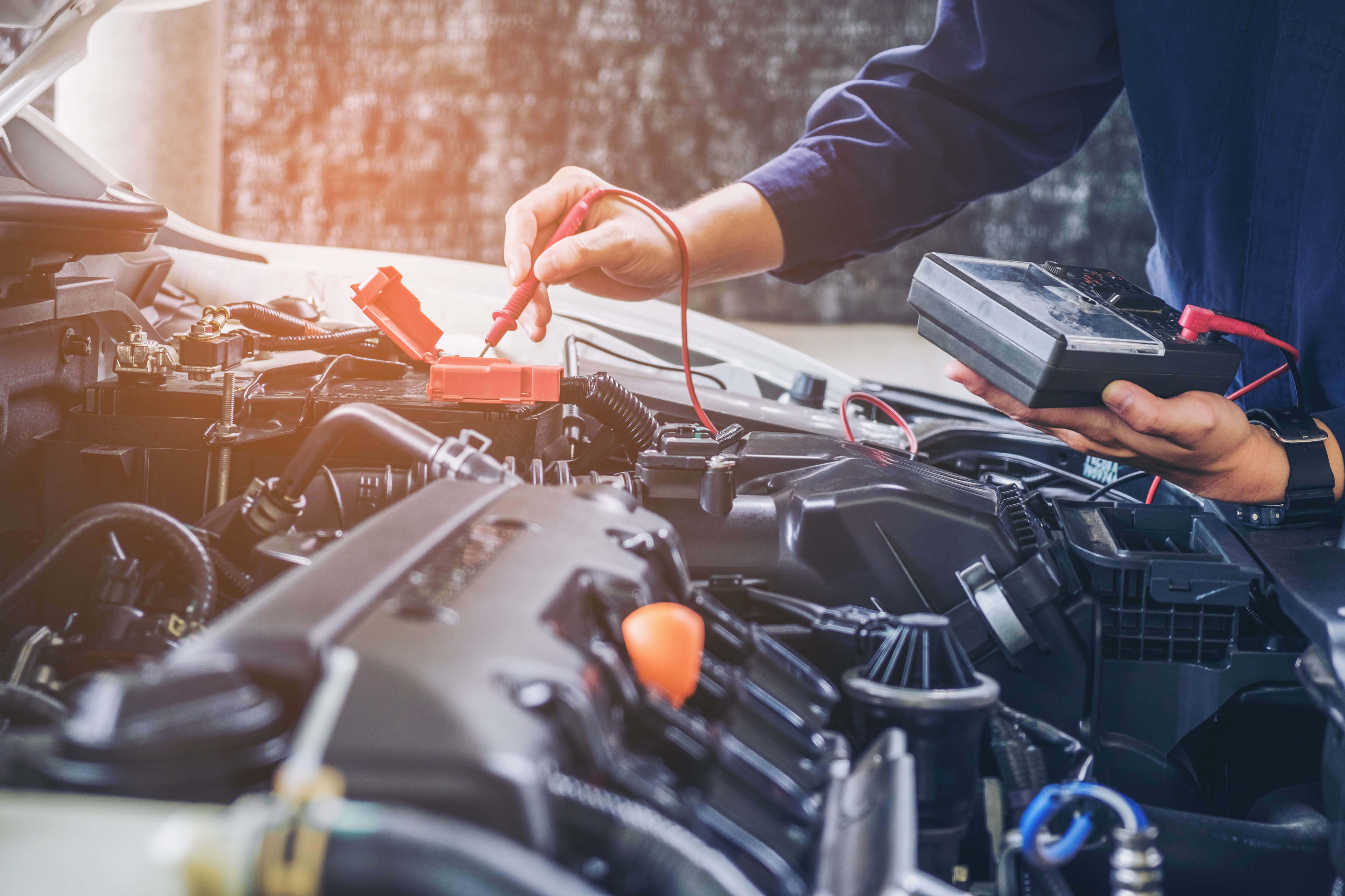 Car Repair 桌布 壁紙 — 一名技師在打開的引擎蓋附近，於溫暖陽光下用萬用電表測試汽車電池。