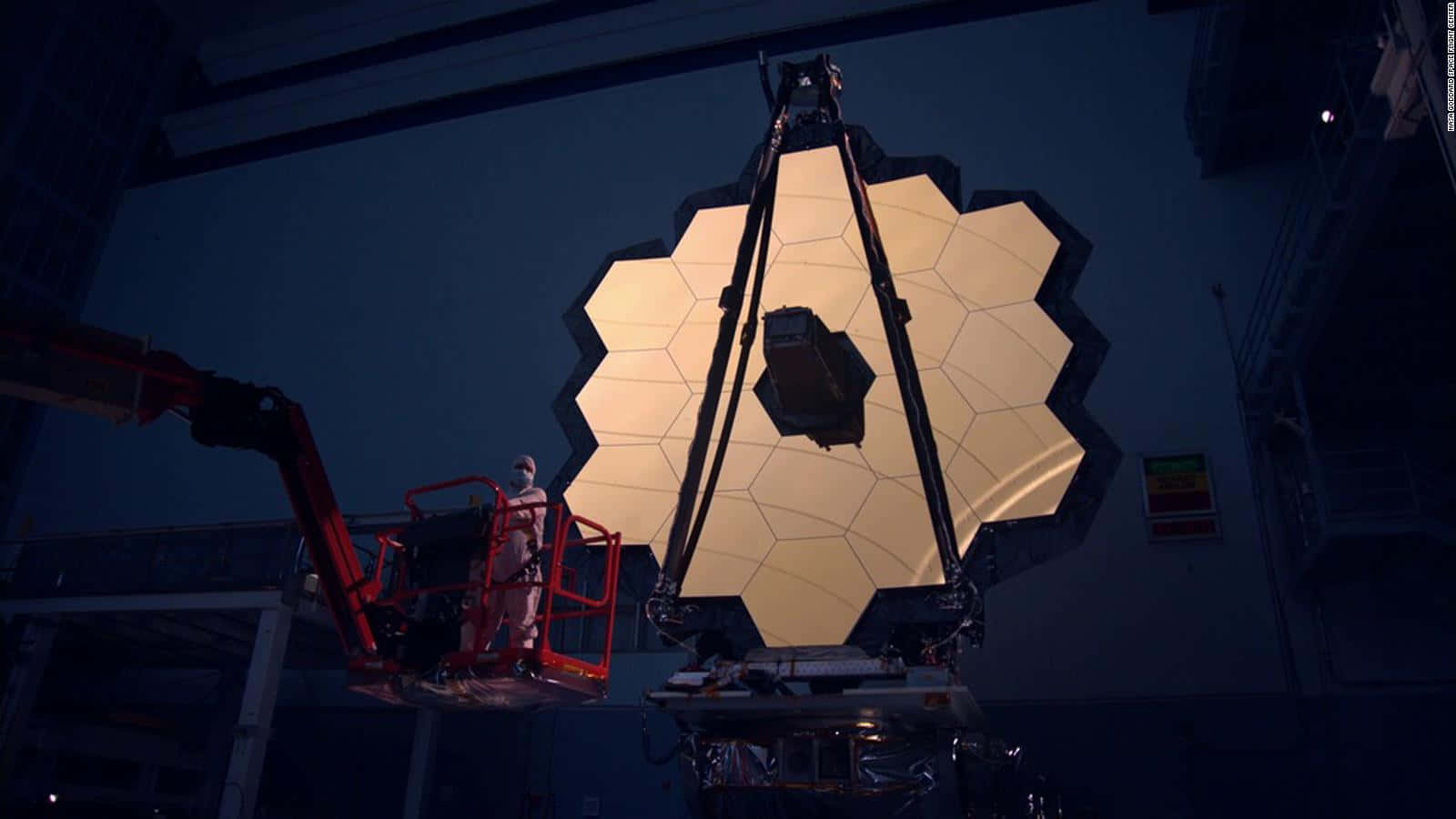 Telescope 桌布 壁紙 — 一名技術人員在紅色升降機上，在昏暗的工業設施內處理一個巨大的六角形金色望遠鏡鏡面。