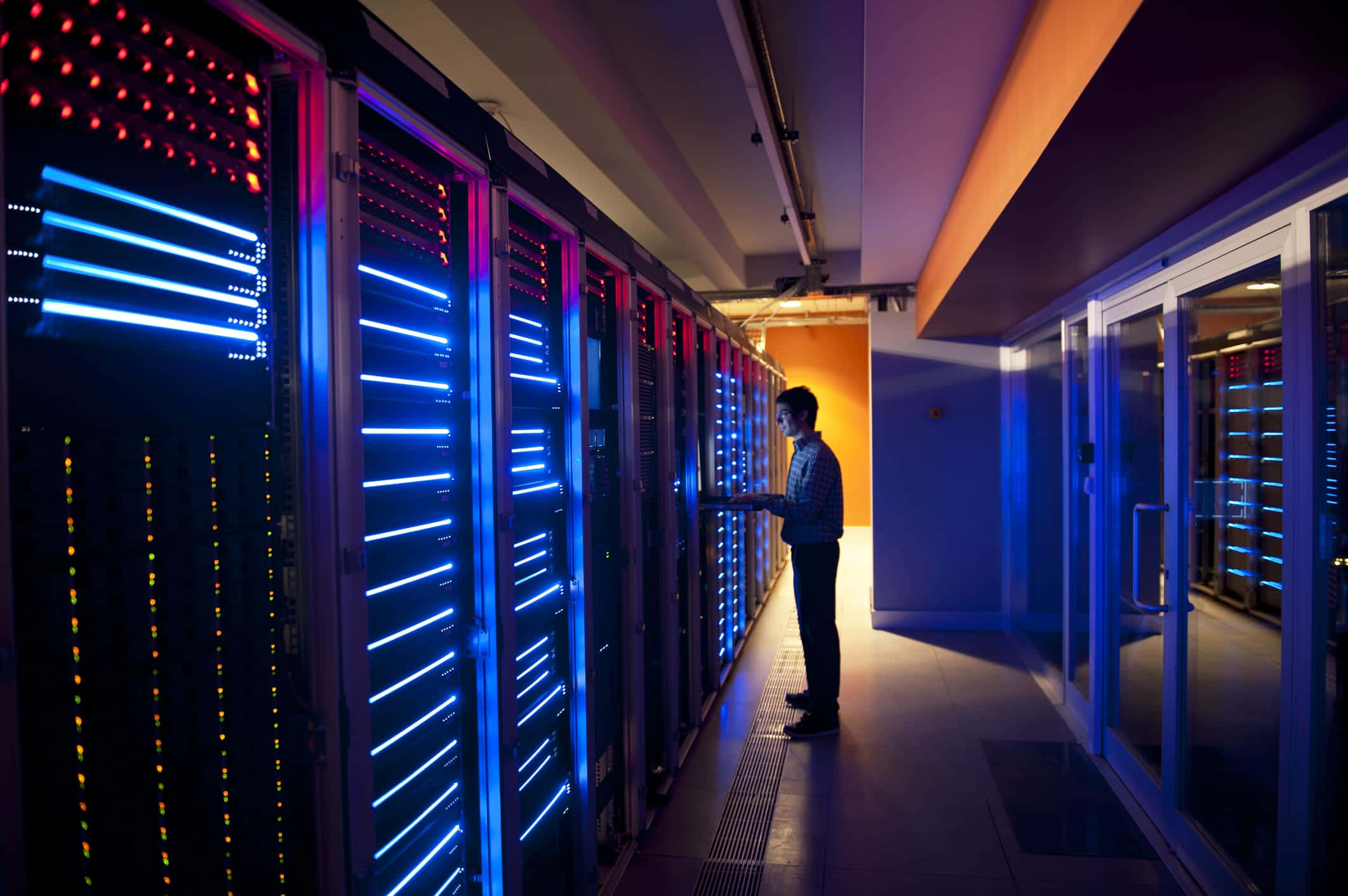 Gaming Servers 桌布 壁紙 — 一名技術人員走過一條高科技數據中心內，兩旁排列著發光藍色和紅色伺服器機架的走廊。