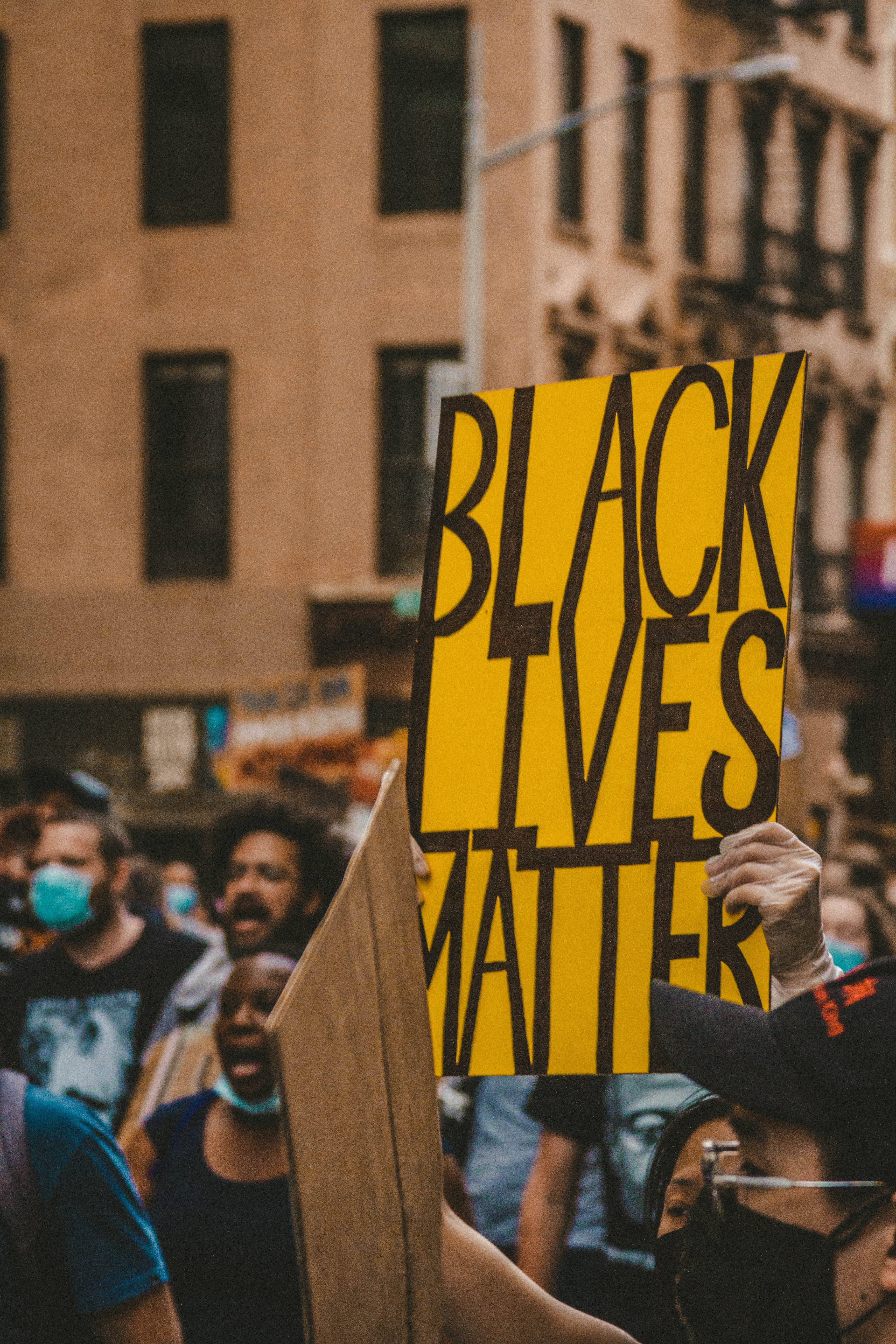 New 桌布 壁紙 — 一名抗議者在城市街道的人群中舉起一個醒目的黃色「BLACK LIVES MATTER」標語牌。