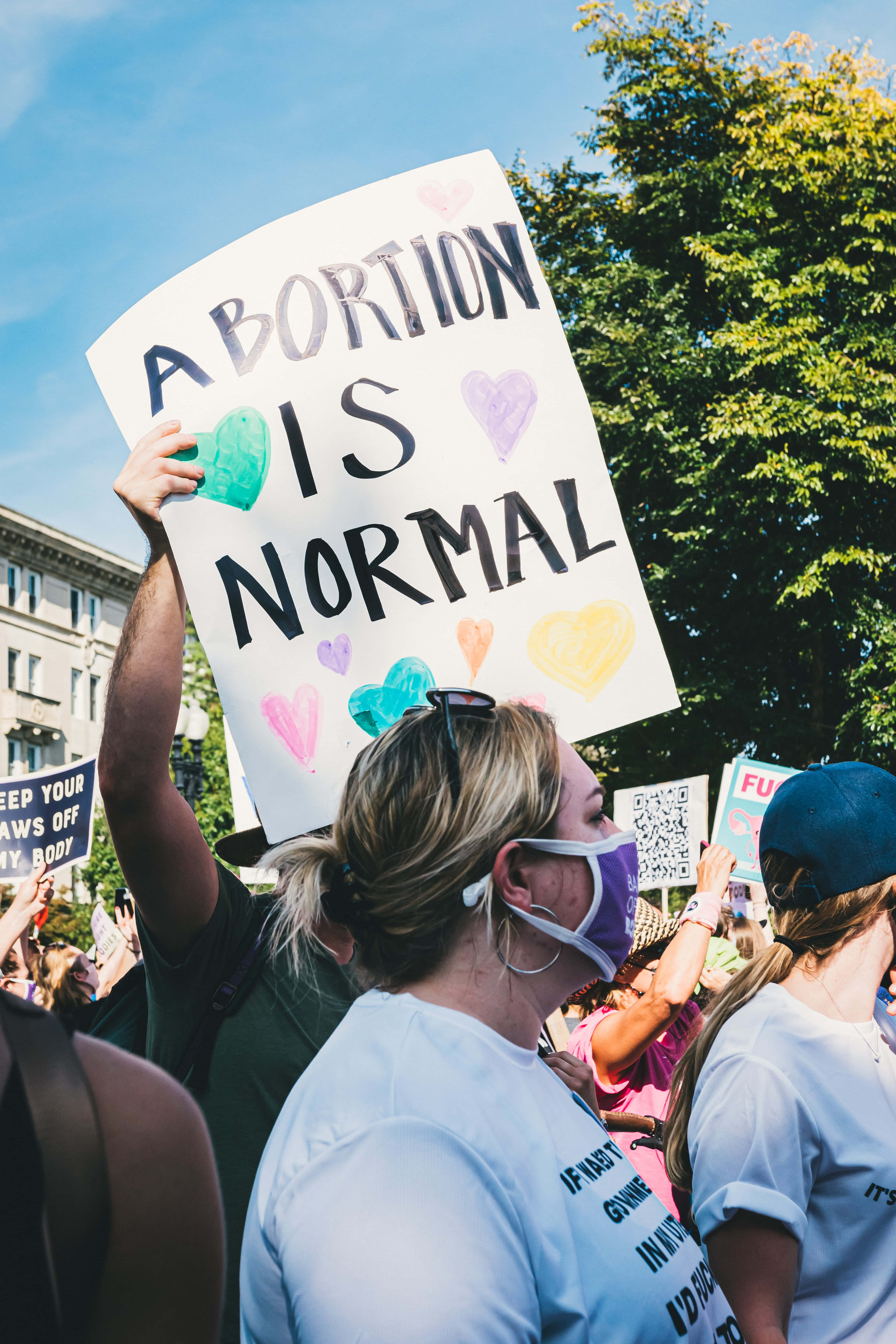 政治 桌布 壁紙 — 一名抗議者在陽光明媚的戶外集會中，手持寫有「ABORTION IS NORMAL」的彩色標語牌，背景有樹木和建築物。