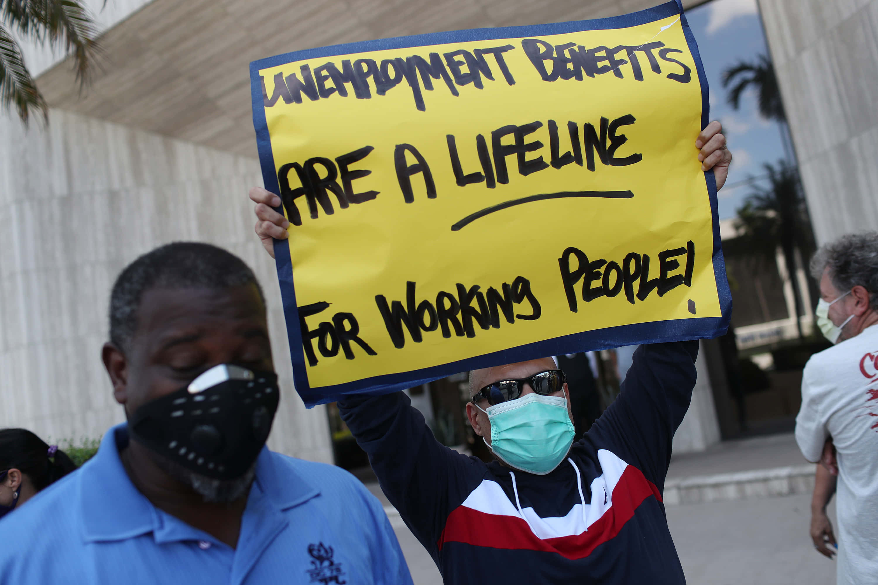 Unemployment 桌布 壁紙 — 一名抗議者手持亮黃色標語牌，上面寫著「失業救濟是勞動人民的生命線！」，同時戴著口罩和太陽眼鏡。
