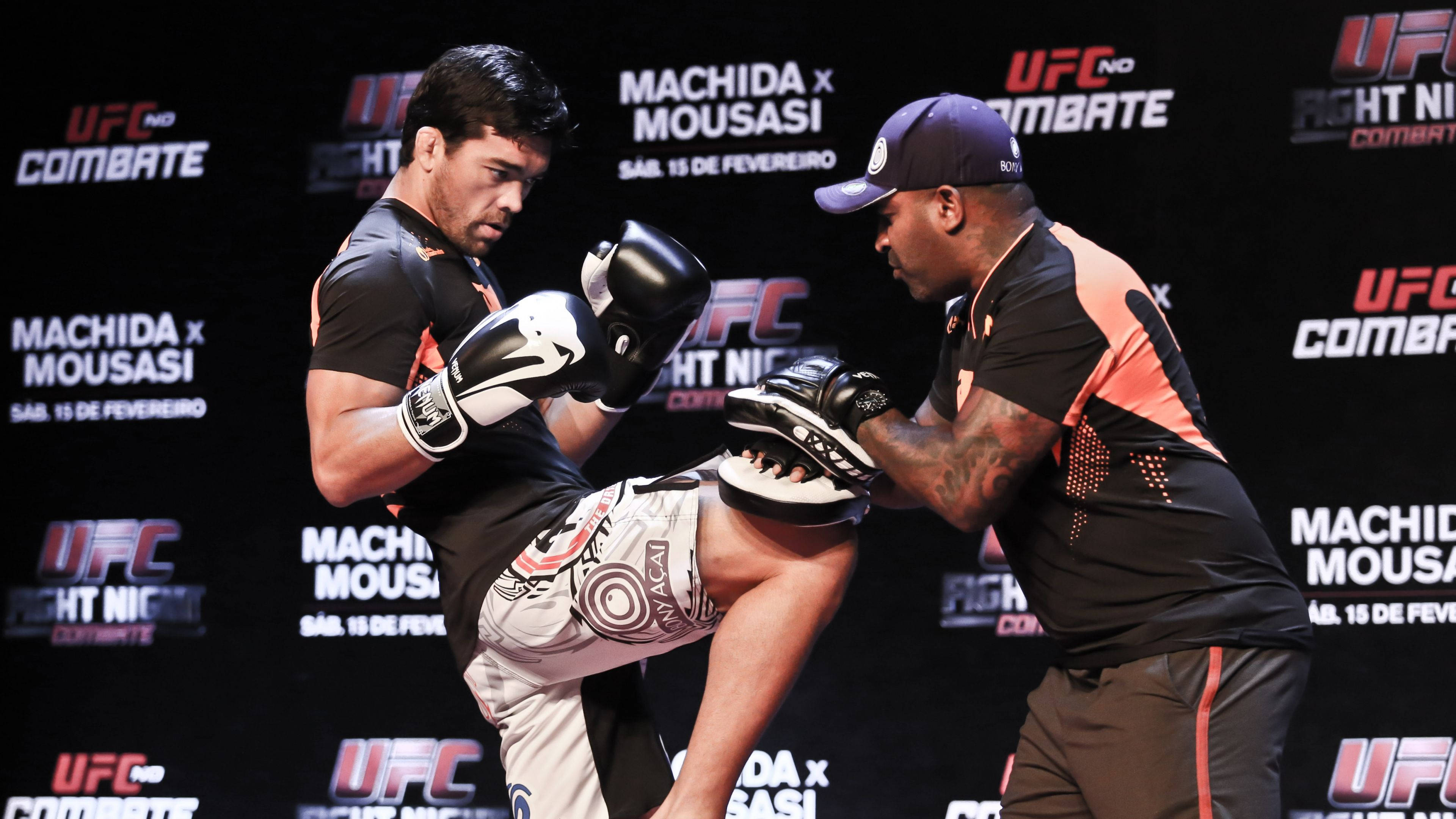 UFC 桌布 壁紙 — 一名拳手在UFC格鬥之夜稱重期間執行高踢，而教練則在品牌背景前拿著護墊。