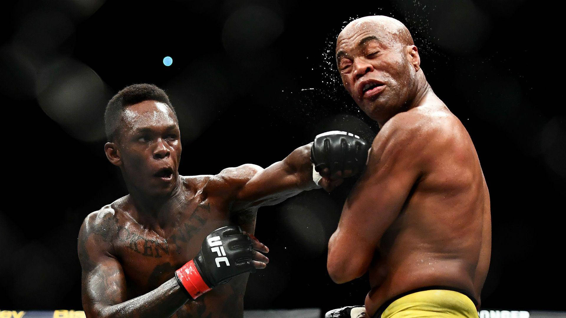 Adesanya 桌布 壁紙 — 一名拳手在燈光明亮的MMA擂台上向對手打出強力一拳，背景是模糊的人群。