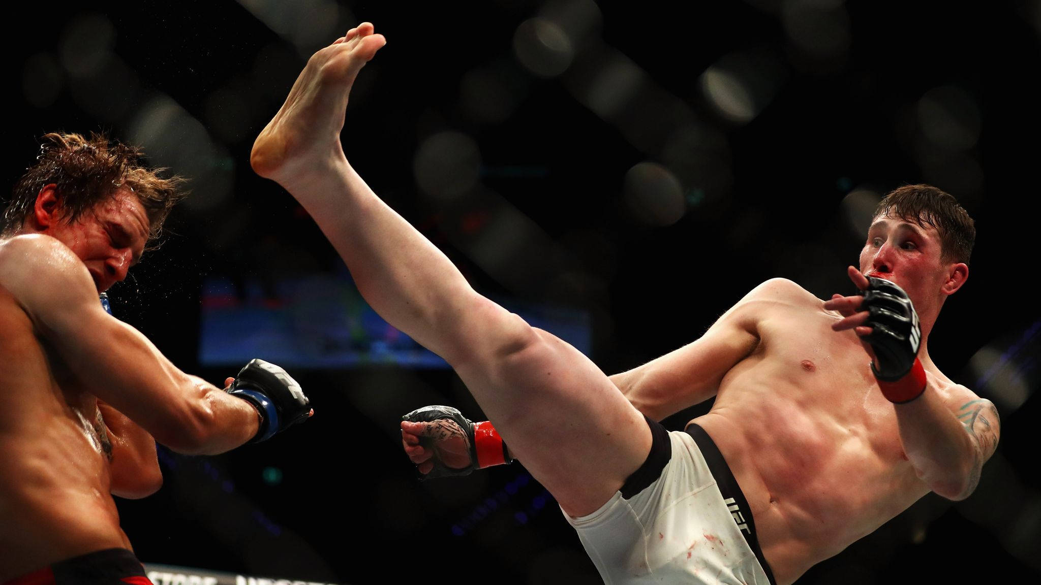 UFC 桌布 壁紙 — 一名拳手在燈光昏暗的MMA鐵籠中向對手使出高踢，背景有模糊的燈光。