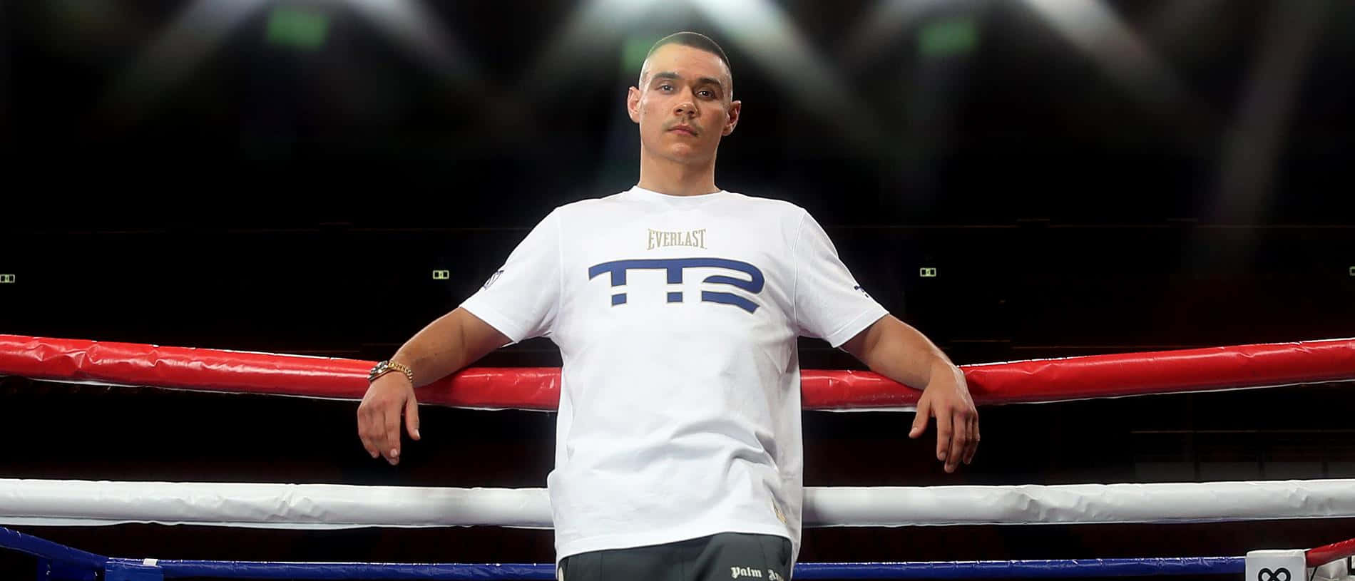 Tim Tszyu 桌布 壁紙 — 一名拳擊手自信地站在一個光線充足、帶有紅色繩索和上方聚光燈的擂台上。