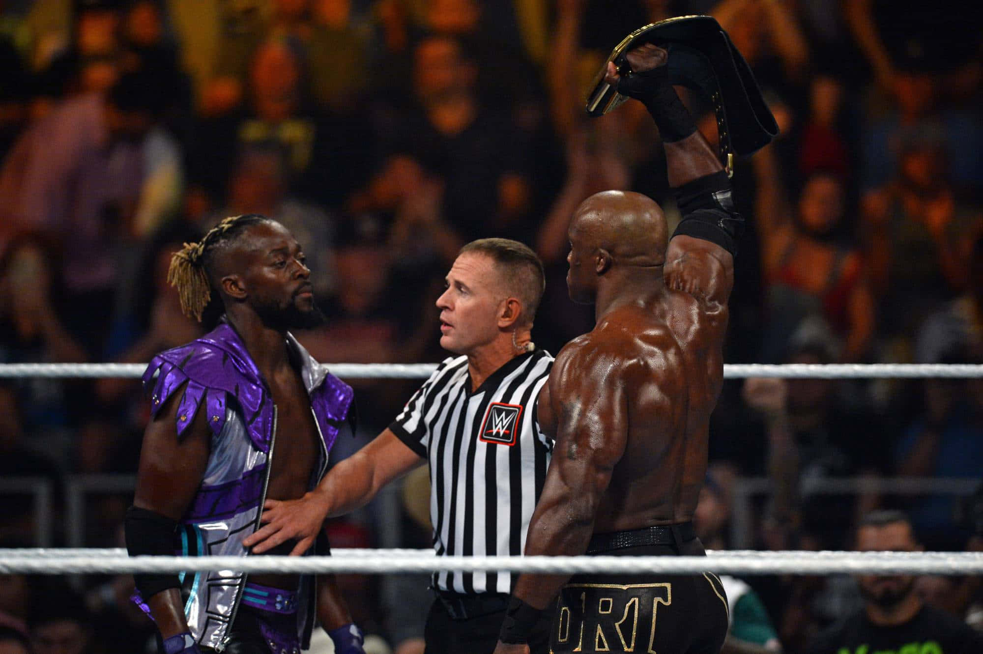 WWE 桌布 壁紙 — 一名摔角手勝利地高舉他的冠軍腰帶，裁判介入他與對手之間，在燈光明亮的擂台上。