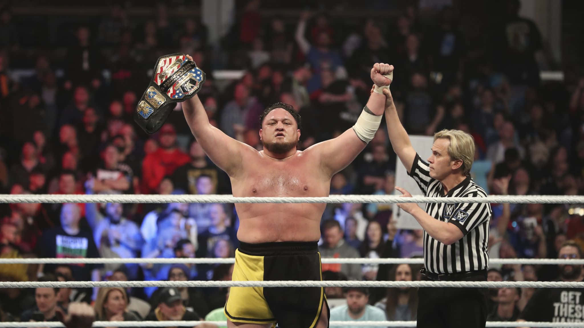 WWE 桌布 壁紙 — 一名摔角手勝利地高舉冠軍腰帶，裁判舉起他的手臂慶祝勝利，擂台內有歡呼的人群。