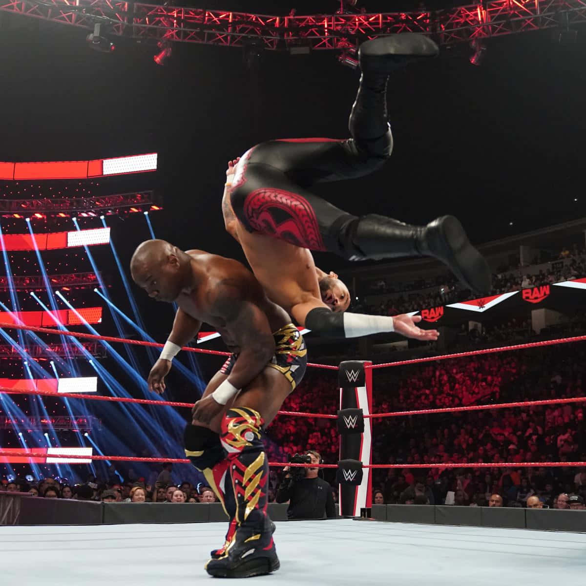 WWE 桌布 壁紙 — 一名摔角手在WWE擂台上跳過另一名摔角手，在明亮的紅藍燈光下，伴隨著咆哮的人群。