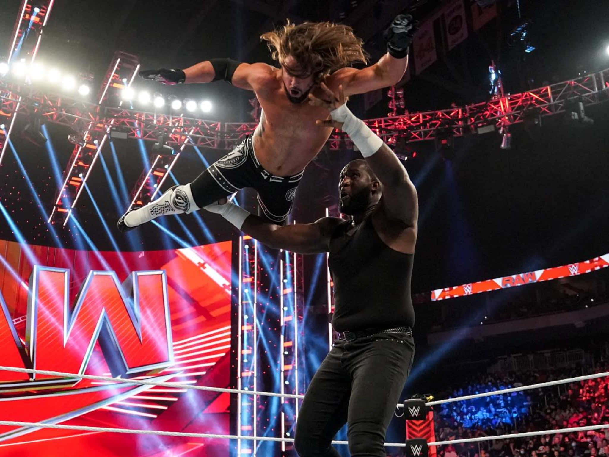 WWE 桌布 壁紙 — 一名摔角手在明亮的舞台燈光下，於摔角擂台上將另一人高舉過頭頂。