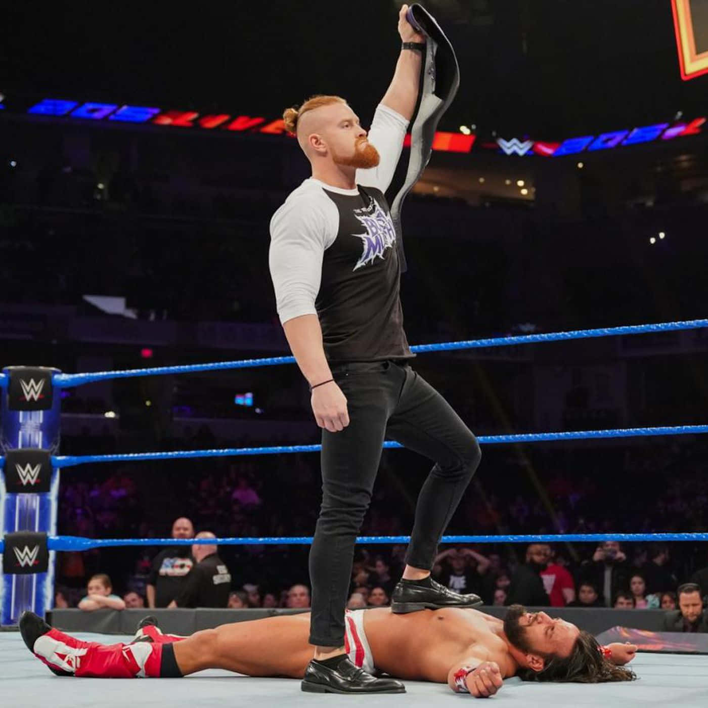 WWE 桌布 壁紙 — 一名摔角手在燈光明亮的擂台上，高舉冠軍腰帶，站在被擊敗的對手身上，慶祝勝利。