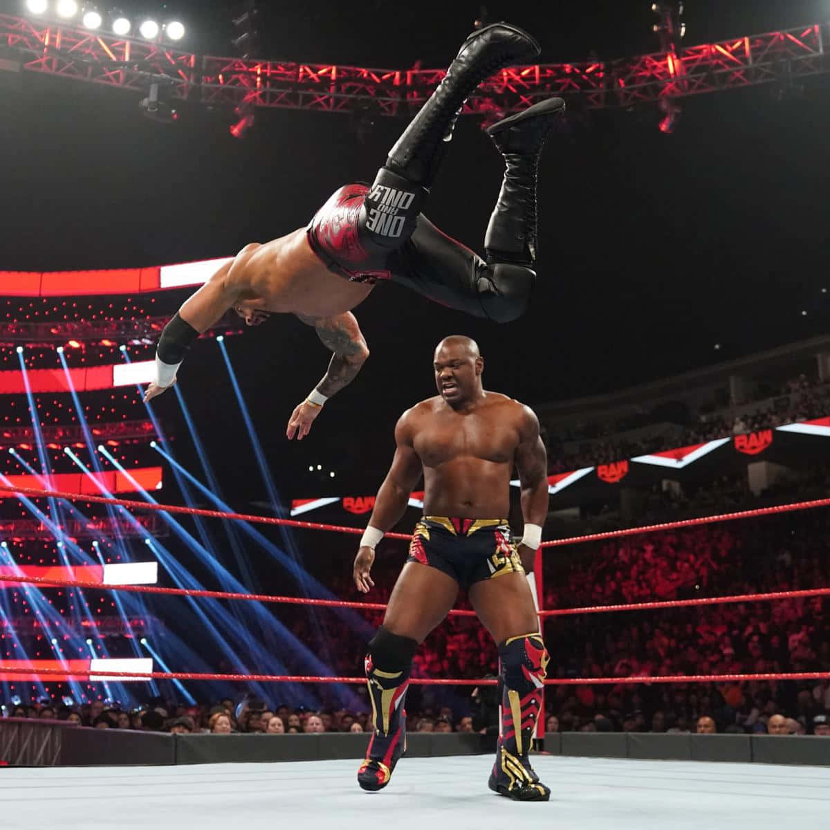 WWE 桌布 壁紙 — 一名摔角手在燈光璀璨、紅光閃爍、觀眾歡呼的擂台上，對另一名摔角手執行高空翻轉。