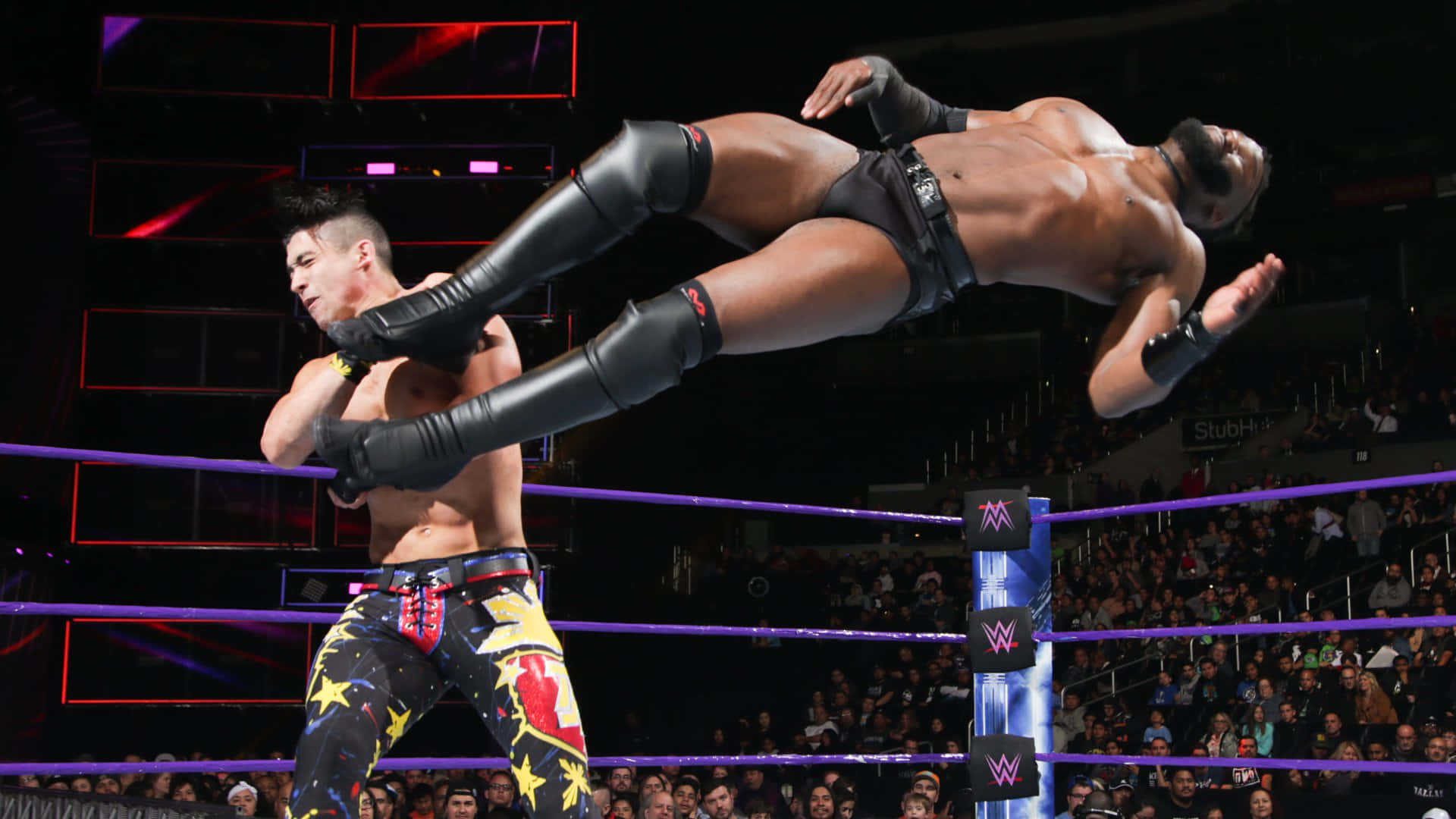 WWE 桌布 壁紙 — 一名摔角手將對手高高舉起，在燈光明亮的擂台上進行戲劇性的空中動作，周圍有歡呼的粉絲。