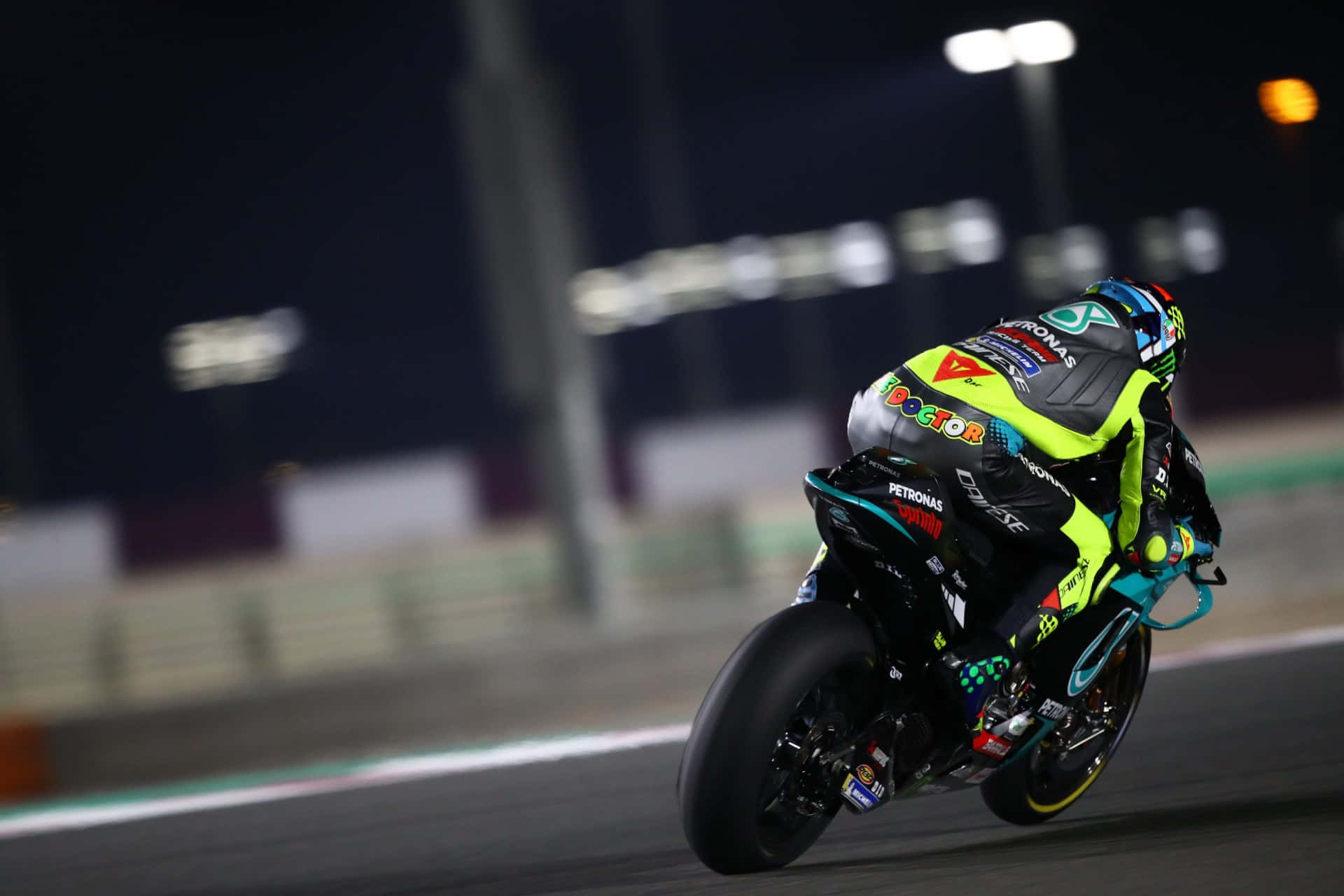 Racing 桌布 壁紙 — 一名摩托車手在夜間燈火通明的賽道上傾斜入一個急彎，背景是模糊的體育場燈光。