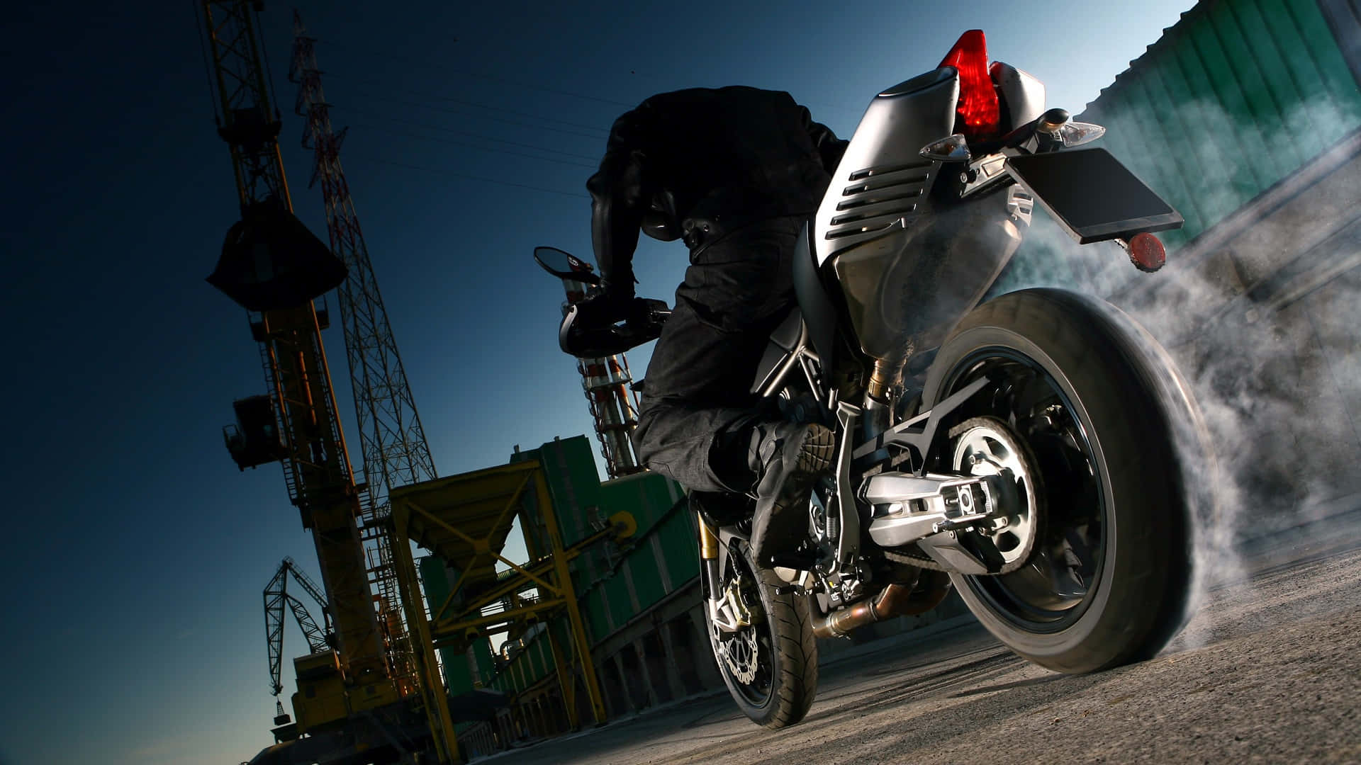 motorbike 桌布 壁紙 — 一名摩托車手在銀色摩托車上傾身進入一個急彎，在黑暗的工業背景下揚起煙霧。