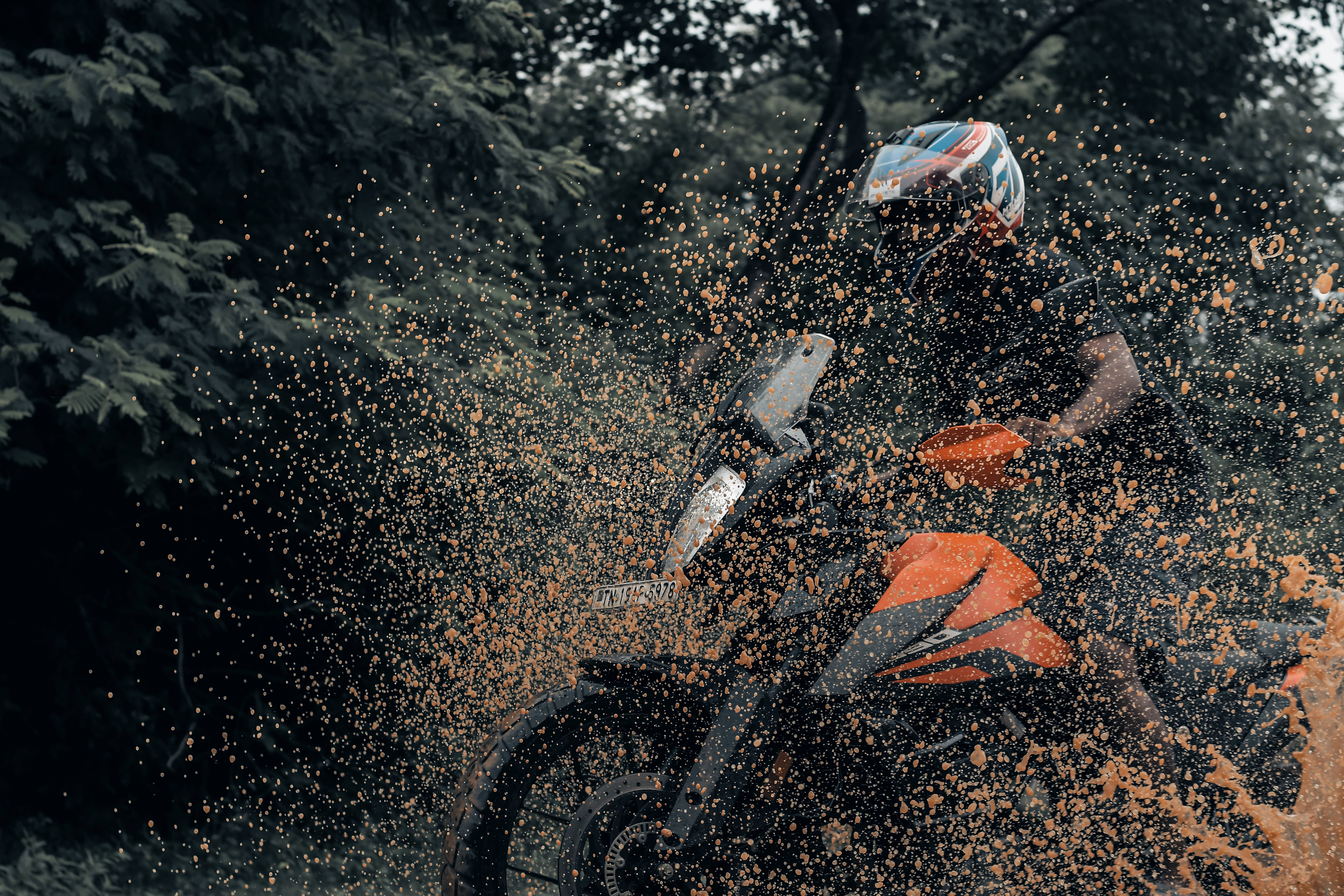 Ktm Rc 390 桌布 壁紙 — 一名摩托車騎士戴著色彩繽紛的安全帽，在森林地區的泥濘地形上行駛，濺起橙色的水花。