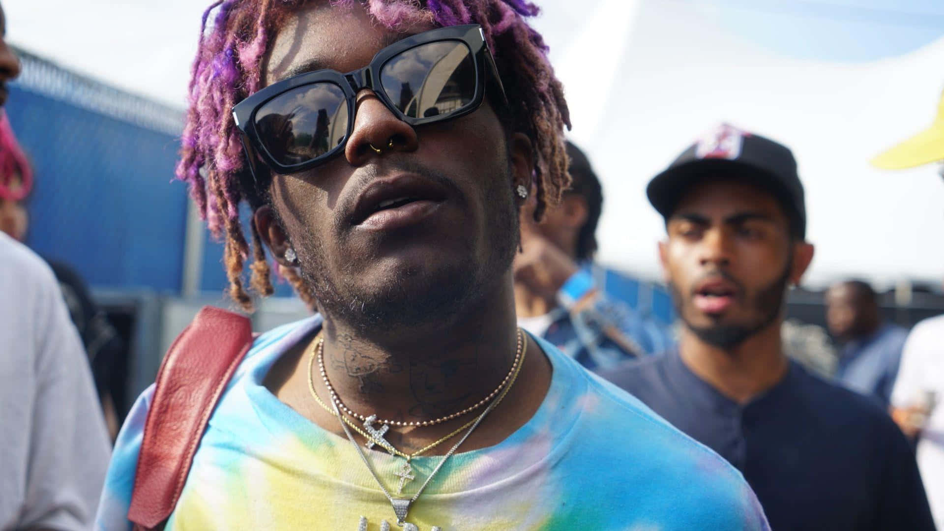 Lil Uzi Album 桌布 壁紙 — 一名擁有充滿活力的紫色和棕色辮子頭及大型黑色太陽眼鏡的男子，穿著紮染襯衫和層疊項鍊，向上仰望。