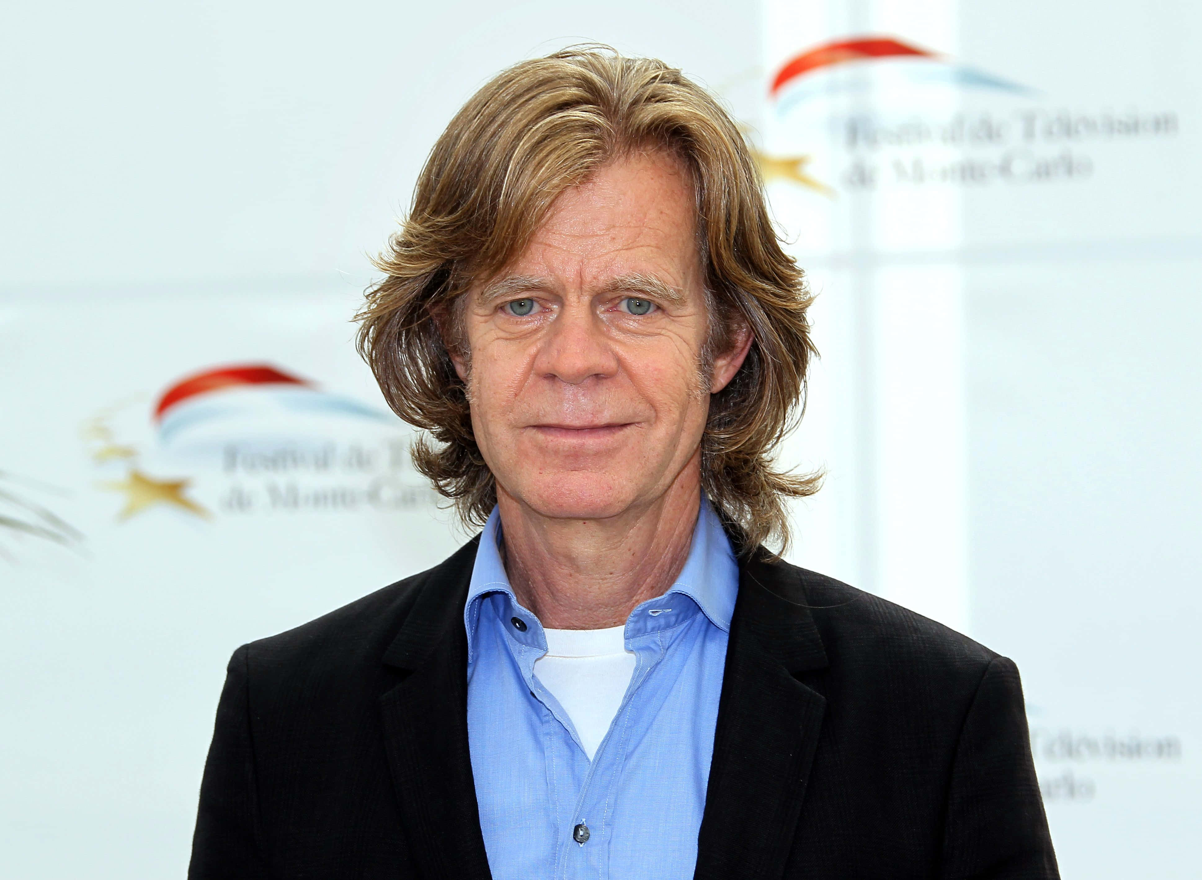 william h. macy 桌布 壁紙 — 一名擁有及肩金髮和藍眼睛的男子，在白色背景上帶有節日標誌的情況下，穿著黑色西裝外套內搭藍色襯衫。