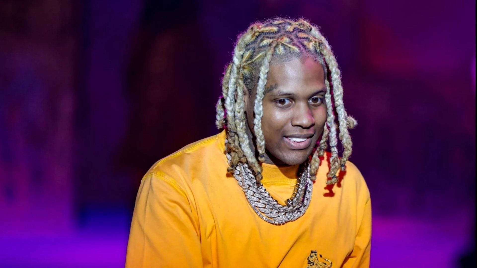 Lil Durk 桌布 壁紙 — 一名擁有彩色辮子頭髮和層疊項鍊的男子，穿著亮橙色襯衫，在紫色燈光背景下微笑。