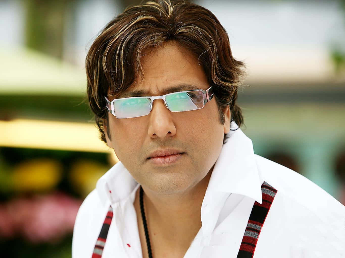 Bollywood Hero 桌布 壁紙 — 一名擁有時尚棕色頭髮和反光眼鏡的男子，在模糊多彩的背景前穿著帶有圖案吊帶的白襯衫。
