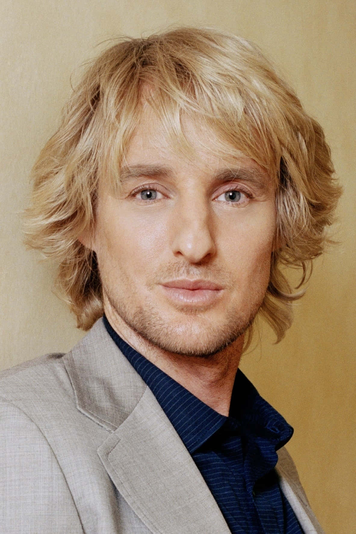 Owen Wilson 桌布 壁紙 — 一名擁有波浪形金髮和淺色眼睛的男士特寫肖像，穿著灰色西裝外套，內搭深藍色襯衫，背景為溫暖的米色。
