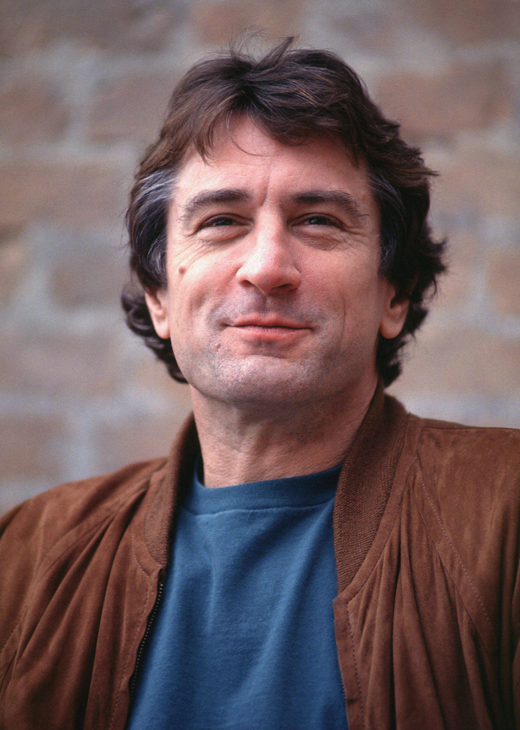 Robert De Niro 桌布 壁紙 — 一名擁有波浪棕色頭髮和溫暖笑容的男子，身穿棕色麂皮外套，內搭藍色T恤，背景是磚牆。