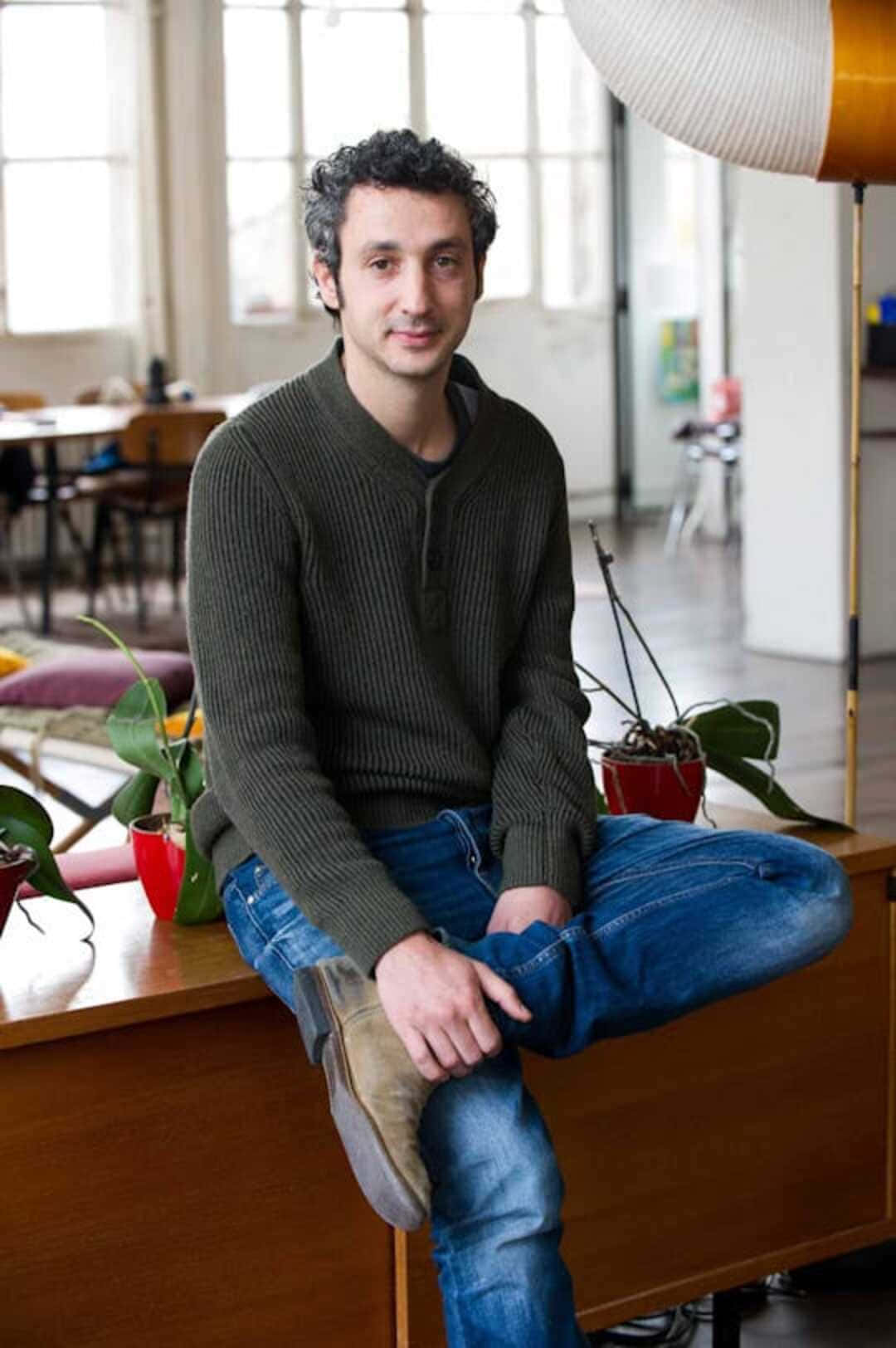 時尚 桌布 壁紙 — 一名擁有深色捲髮的男子，隨意地坐在一個光線充足、通風良好的房間內的木桌上，房間有大窗戶和現代裝飾。