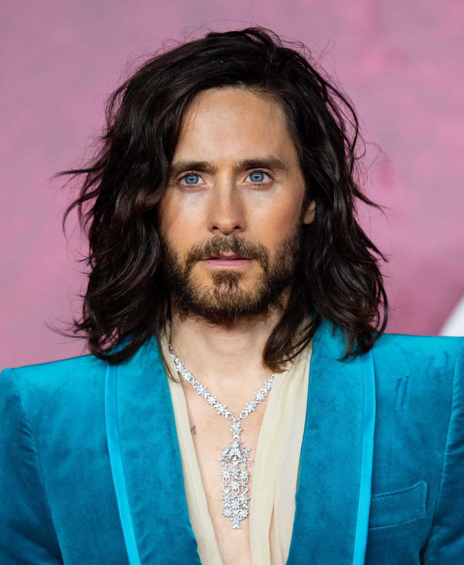 jared leto 桌布 壁紙 — 一名擁有深色長髮和藍色眼睛的男子，在柔和的粉紅色背景前，穿著鮮豔的藍色天鵝絨西裝外套和閃閃發光的鑽石項鍊。