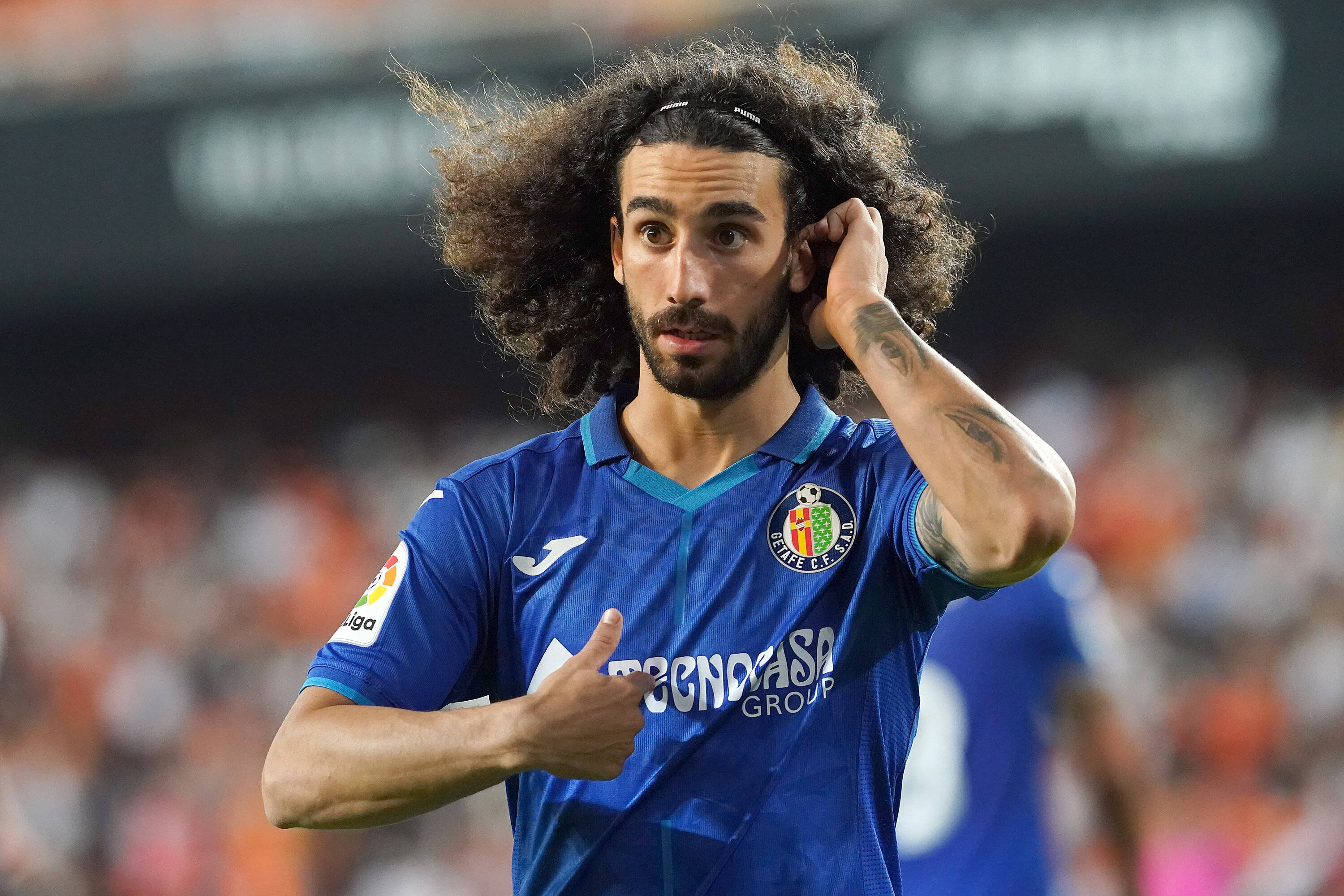 Football Players 桌布 壁紙 — 一名擁有狂野捲髮和刺青的足球運動員在明亮燈光下的體育館豎起大拇指。