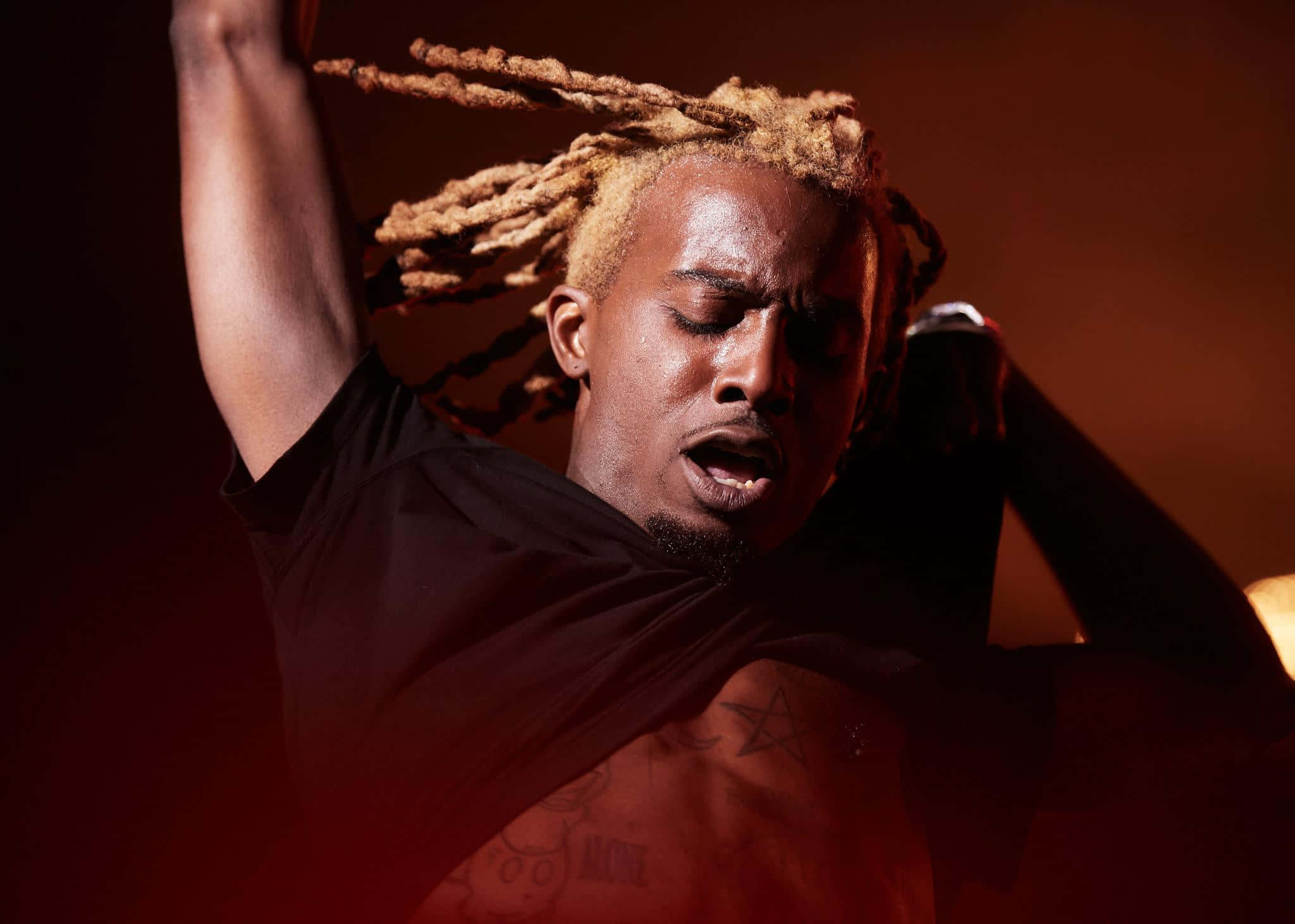Playboi Carti 桌布 壁紙 — 一名擁有金色辮子辮的男人在溫暖的舞台燈光下熱情歌唱，胸前的刺青清晰可見。