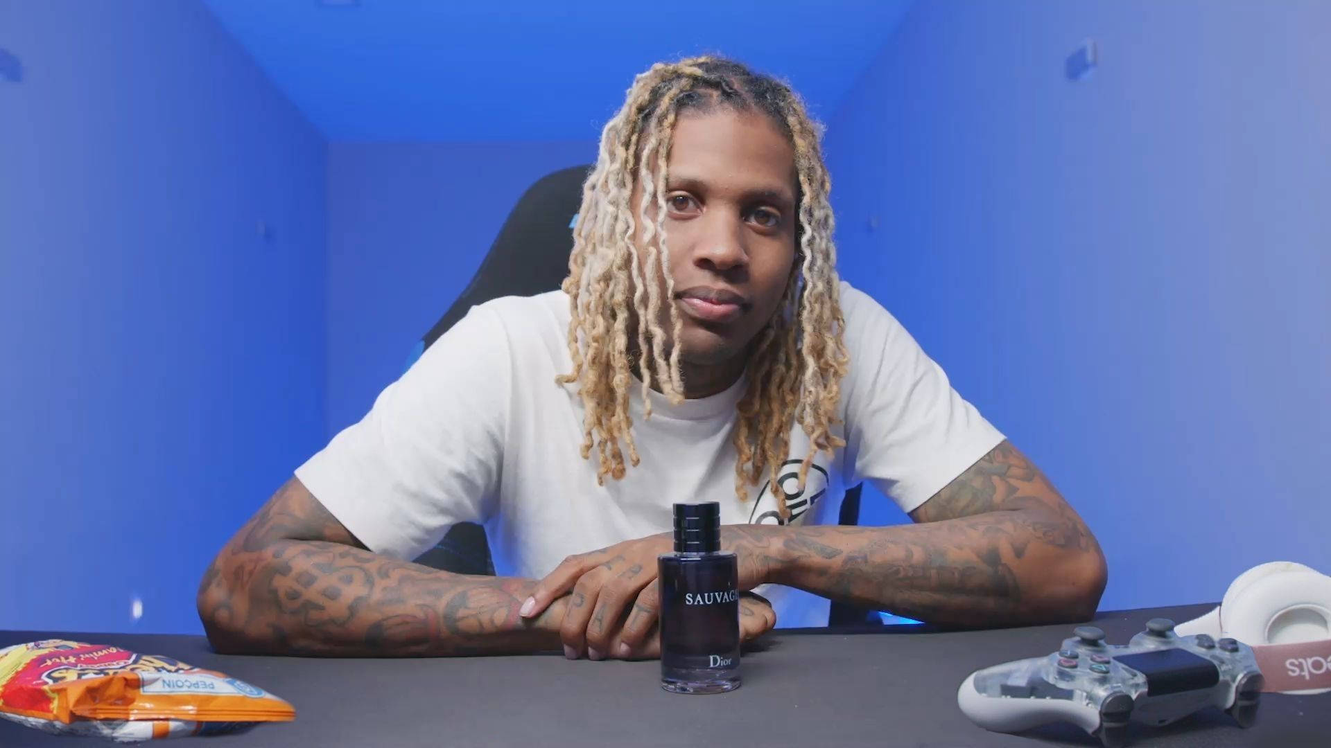 Lil Durk 桌布 壁紙 — 一名擁有金色長髮辮和刺青的男子坐在藍色房間的書桌前，手持香水瓶，附近有一個遊戲控制器。