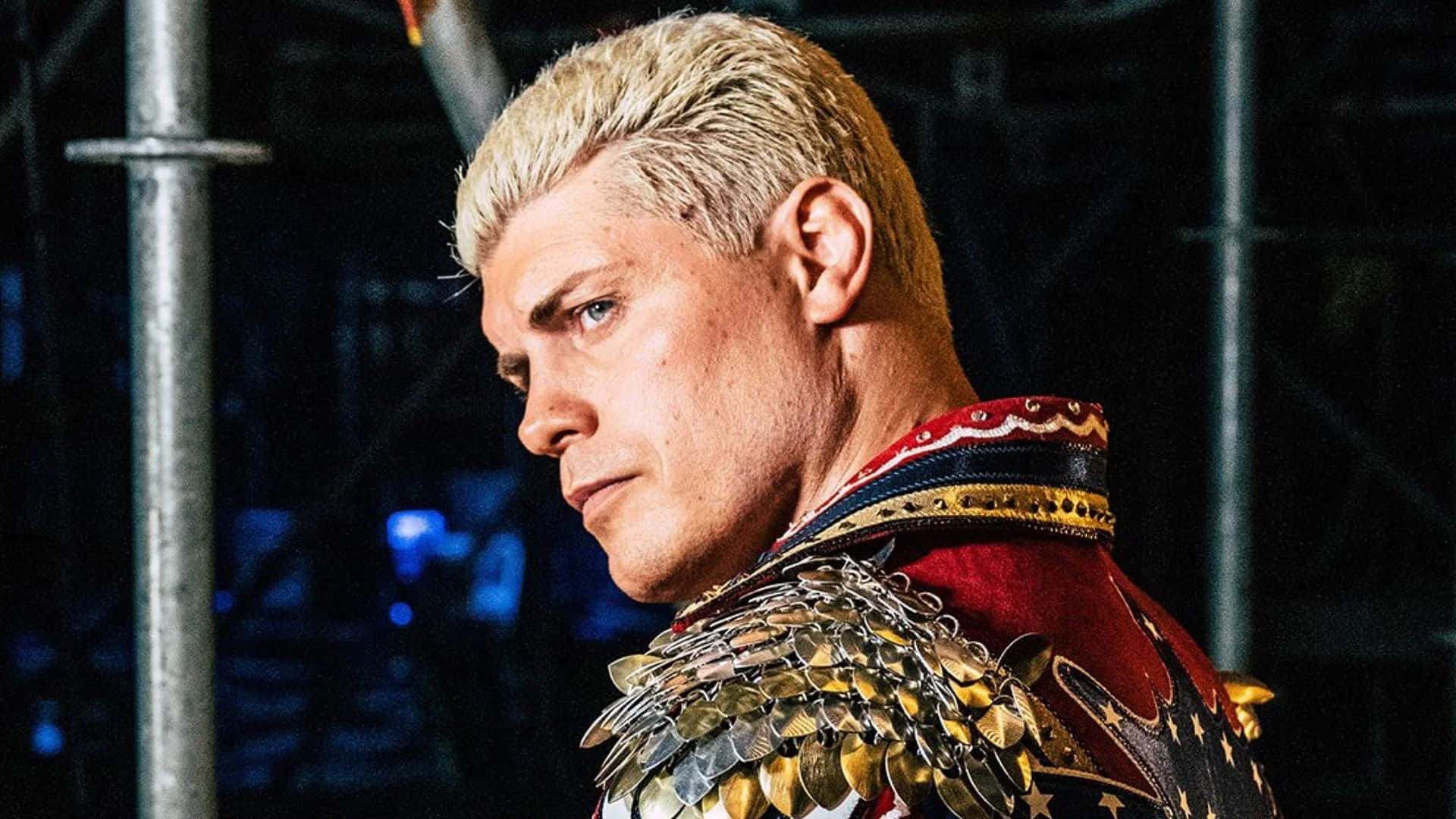 WWE 桌布 壁紙 — 一名擁有鉑金色頭髮和銳利眼神的男子，在黑暗的工業背景下穿著華麗的紅金黑色盔甲。