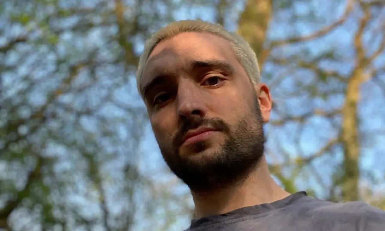 Tom Parker 桌布 壁紙 — 一名擁有鉑金色頭髮和鬍鬚的男子，在模糊的樹木和藍天背景下直視鏡頭。